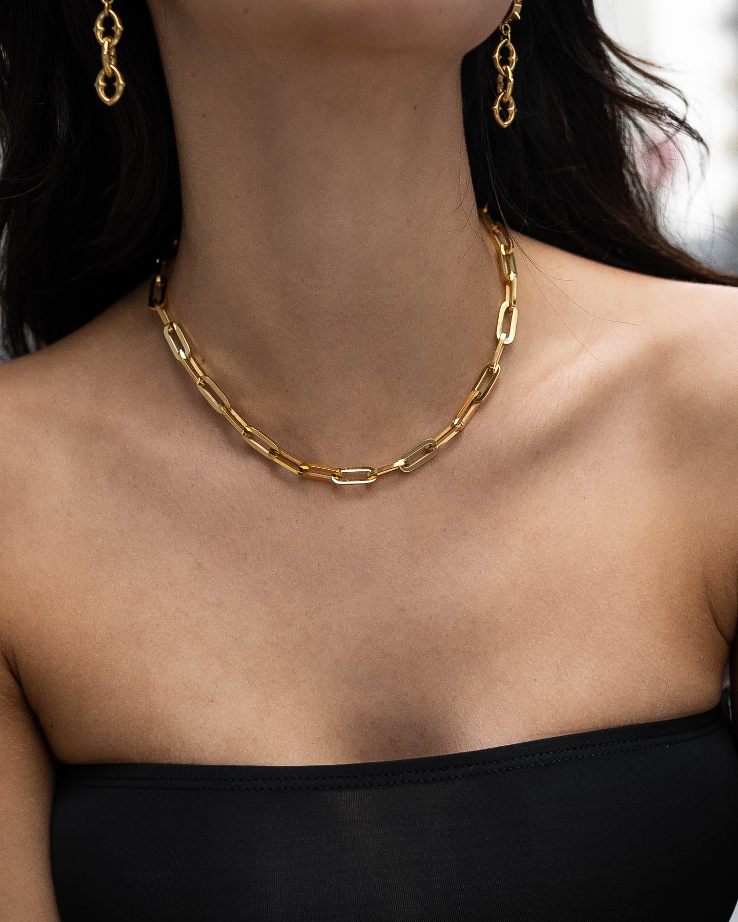A woman in a strapless black top wears the Paperclip Chain (Gold), an 18k gold, non-tarnish necklace, along with matching chain earrings. The bold accessories highlight her neck and collarbone.