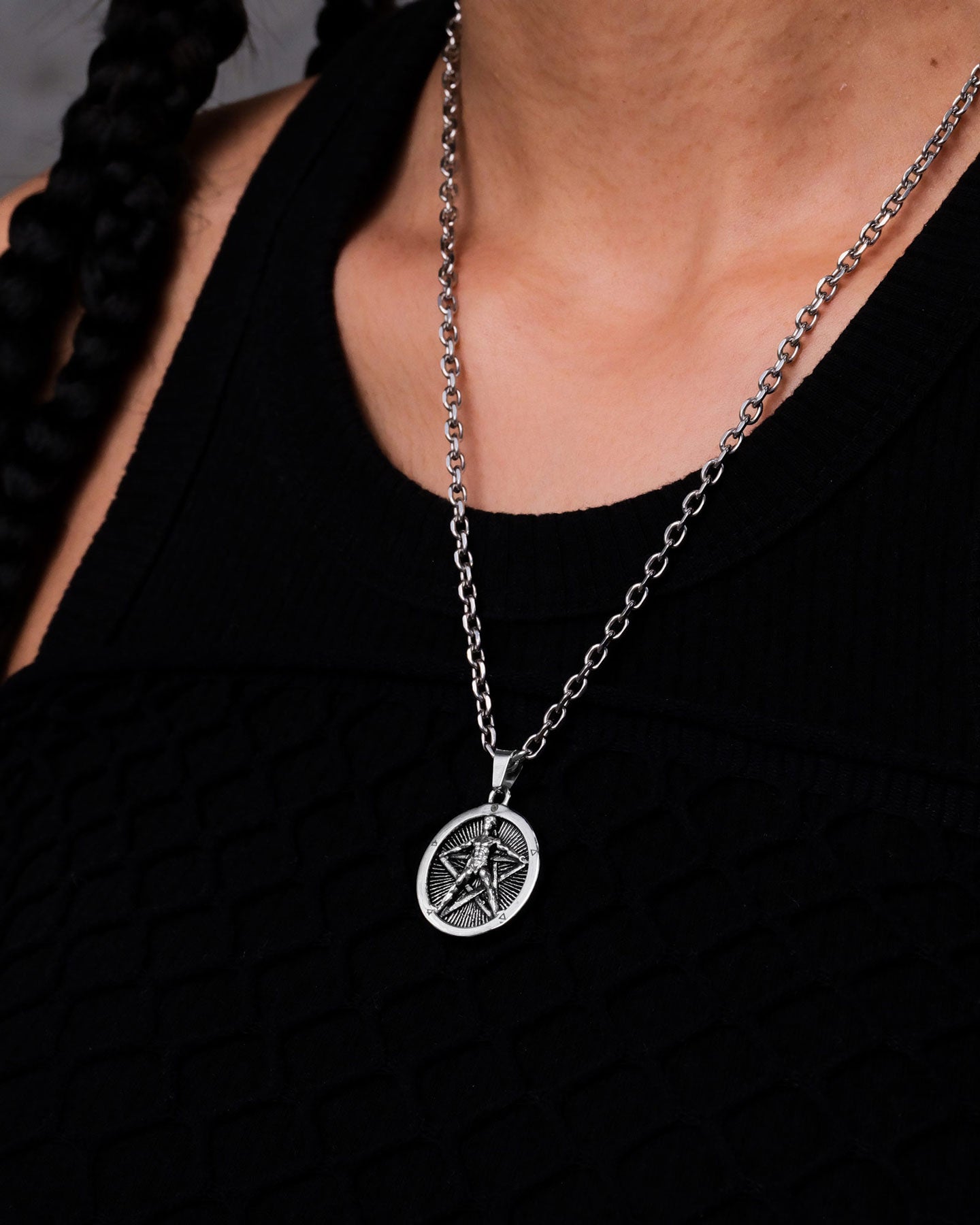 A person with braided hair wears a black textured top and "The Star of the Magi" Pendant—a silver stainless steel necklace with a round, engraved alchemist symbol—shown in focus on their upper chest.