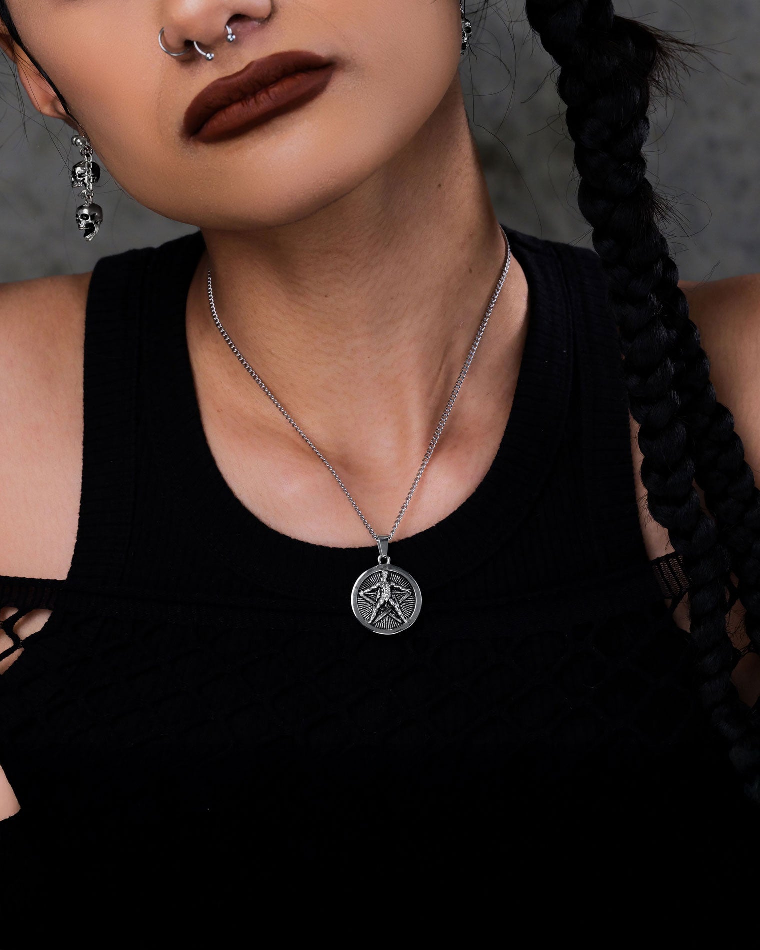 A person with dark lipstick, a nose ring, and skull earrings poses in a black top. Their braided hair highlights "The Star of the Magi" Pendant—a stainless steel necklace featuring a round pentagram inspired by Agrippa.