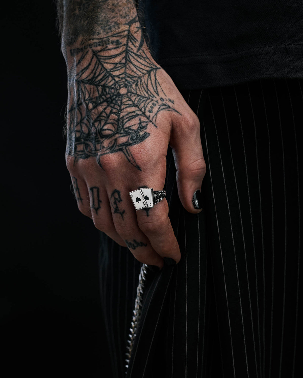 A tattooed hand with a spider web design and black nail polish, adorned with Statement Collective’s Pocket Aces Ring—a macabre jewellery piece featuring two dice—rests against black pinstriped pants, showcasing stainless steel accents and finger tattoos.