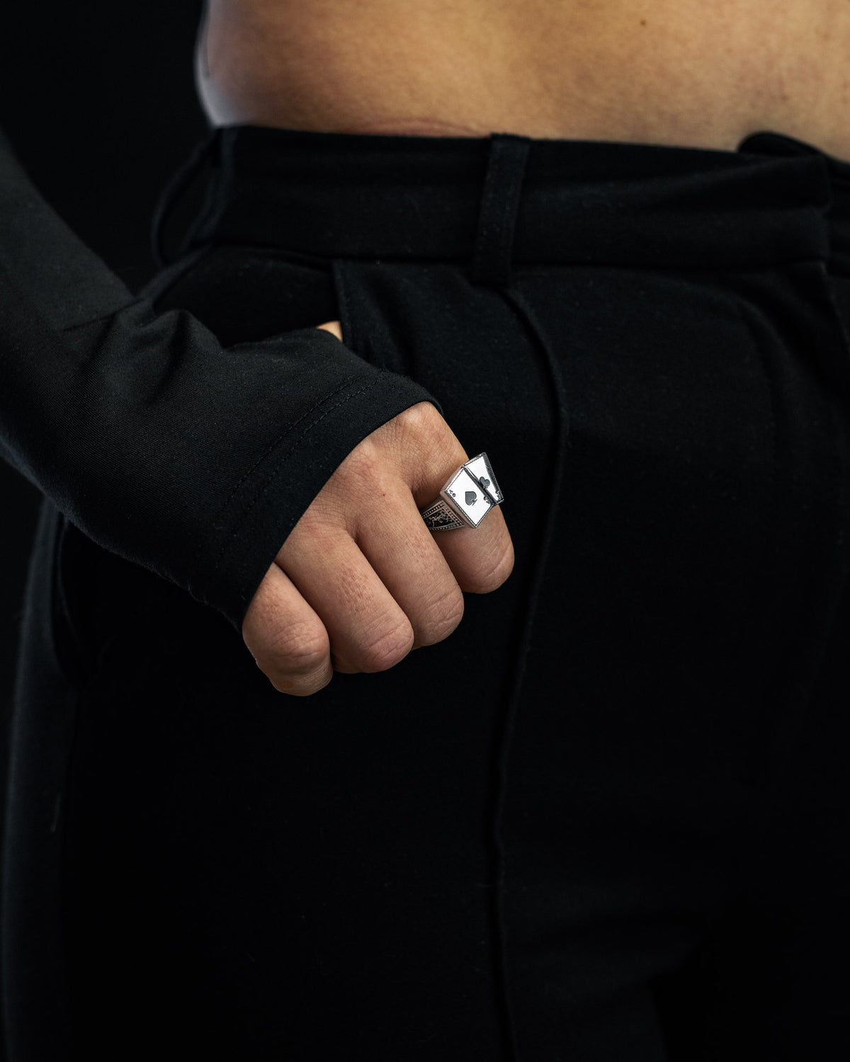 A person dressed in black pants and a long-sleeved black top stands with their fist at their waist, showcasing the Statement Collective Pocket Aces Ring—a bold, chunky stainless steel geometric piece evocative of macabre jewellery.