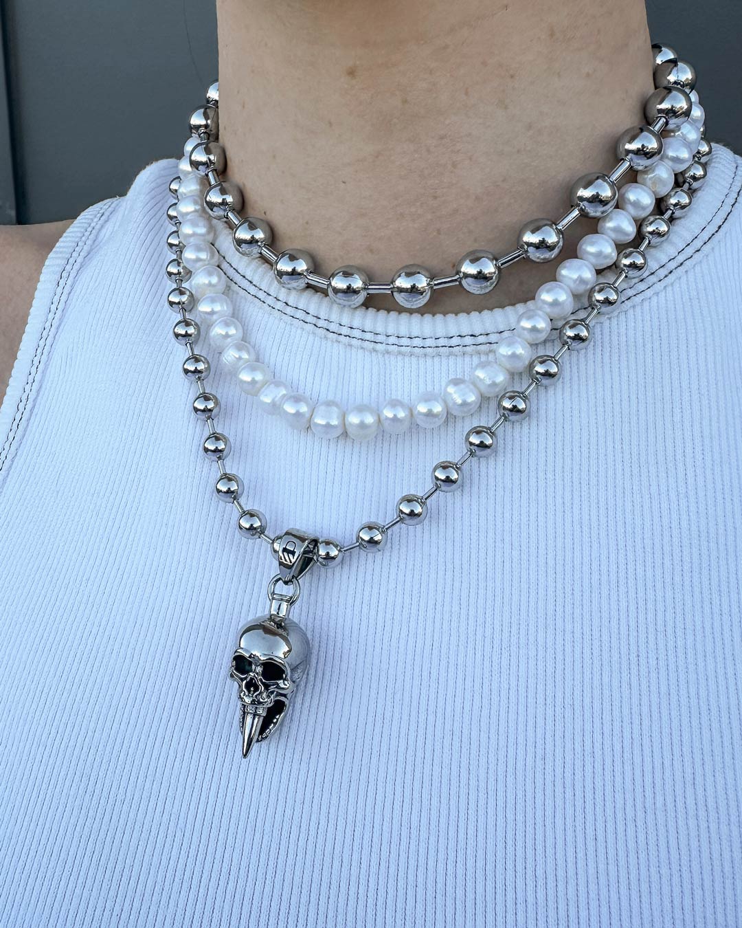 Someone wearing the Precious Death Stack: a layered necklace combo featuring silver beads, pearls, and a bullet skull necklace with a metallic skull pendant with a long, pointed chin—styled with a white ribbed tank top.