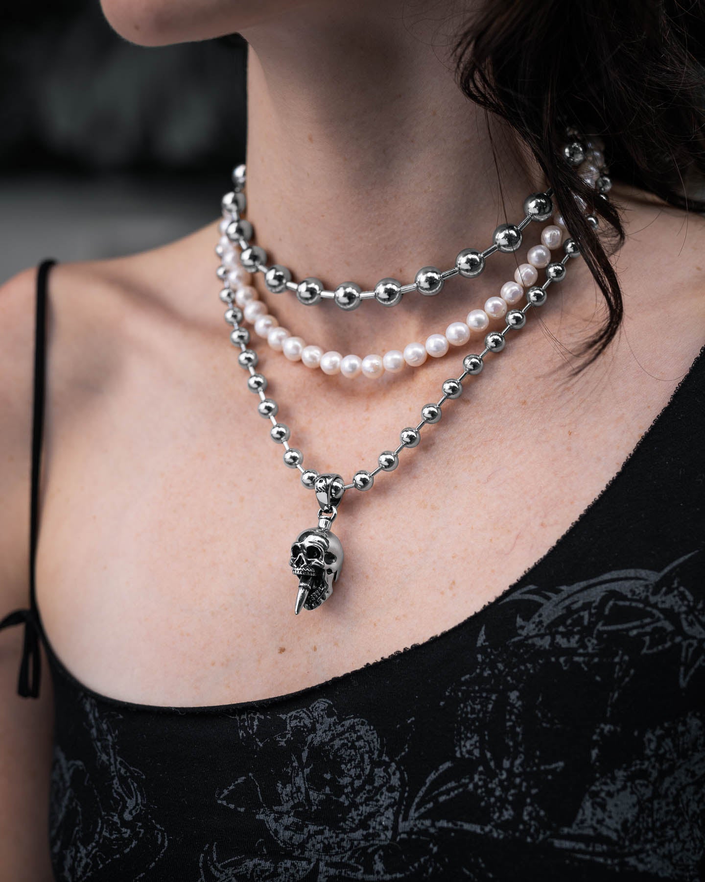 A person wears the Precious Death Stack—a bold bullet skull necklace, a white pearl strand, and chunky silver beads—paired with a black floral-patterned top.
