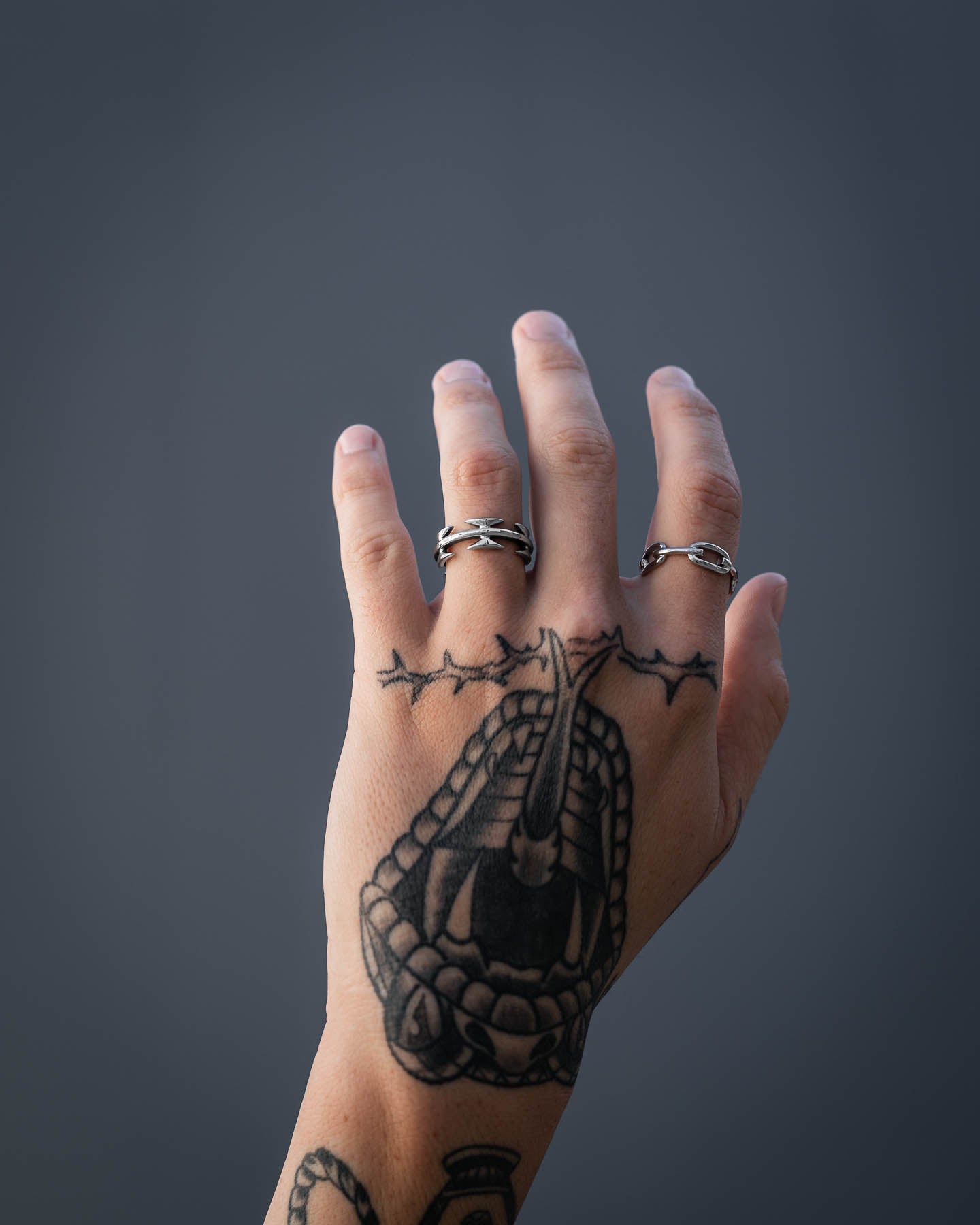 A tattooed left hand with two silver rings, one being the Razor Wire Ring on the middle finger, is held up against a plain dark gray background. The tattoos feature a barbed wire design across the knuckles.