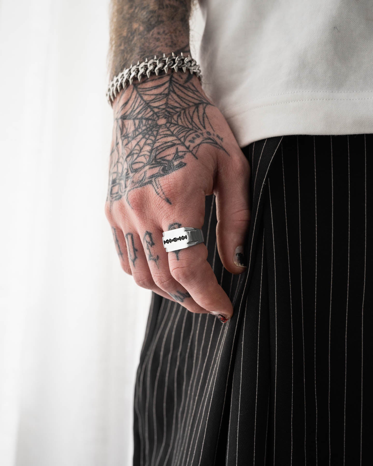 A close-up of a tattooed hand with black nail polish wearing the Razor Blade Signet Ring and a chunky bracelet. Paired with a white shirt and black pinstriped pants, this look showcases a love for bold punk jewelry.