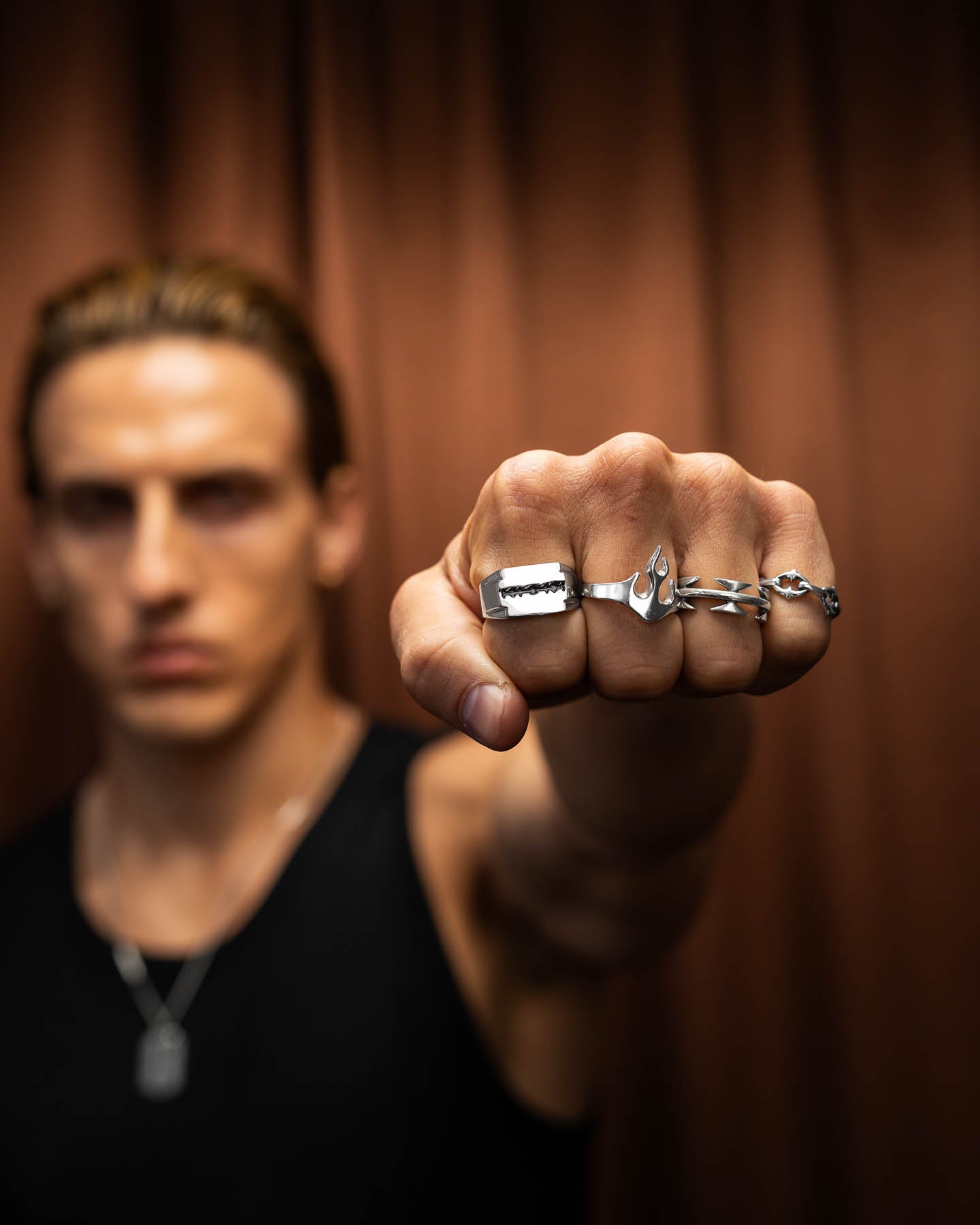 A person in a black tank top and necklace extends a clenched fist with five silver punk rings, featuring the Razor Blade Signet Ring. Brown curtains blur in the background; their face is out of focus.