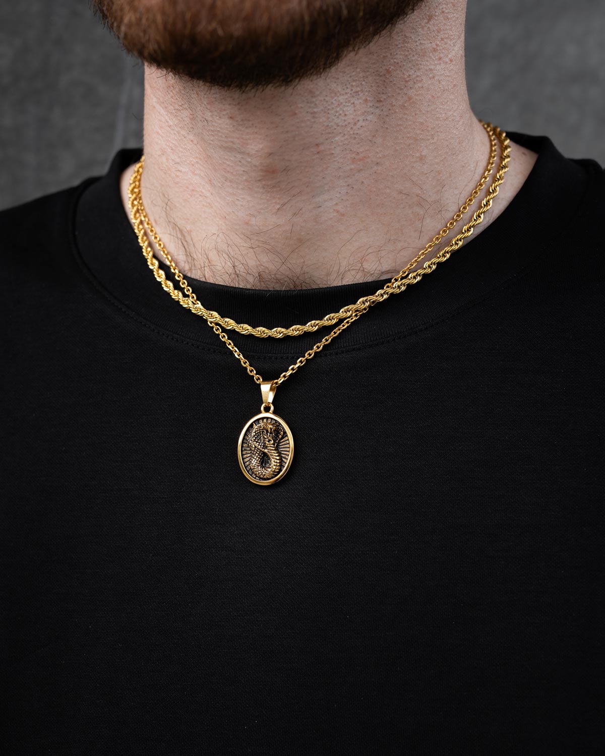 A person wears the Statement Collective Rope Set (Custom Pendant Choice) in gold—a set of three layered gold necklaces including an 18k gold rope chain and a custom oval pendant—over a black shirt, with a neutral gray background.