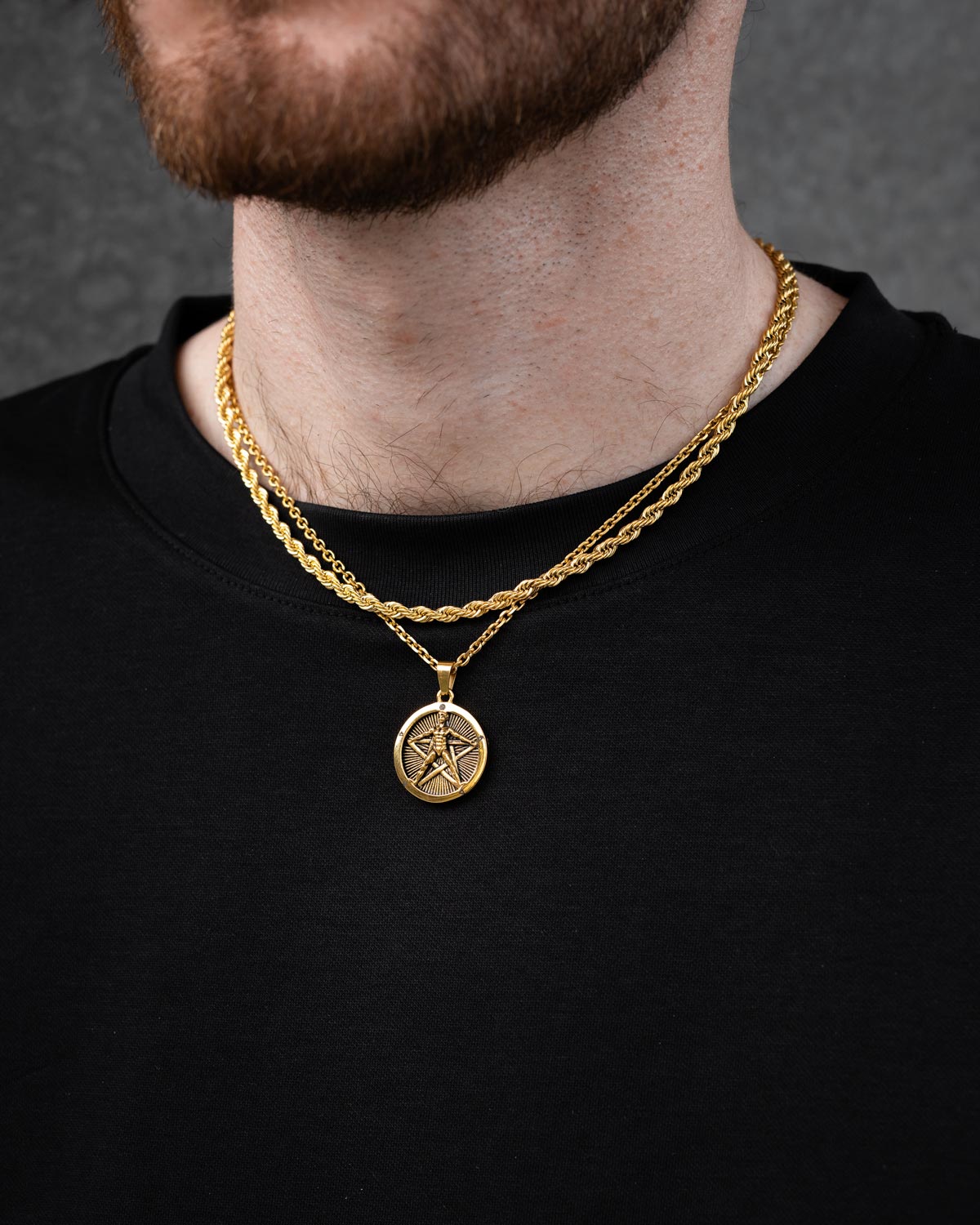 A man in a black shirt wears the Statement Collective Rope Set (Custom Pendant Choice) (Gold), including a gold chain and a round pendant with a five-pointed star in a circle. His brown-bearded face is partially visible.