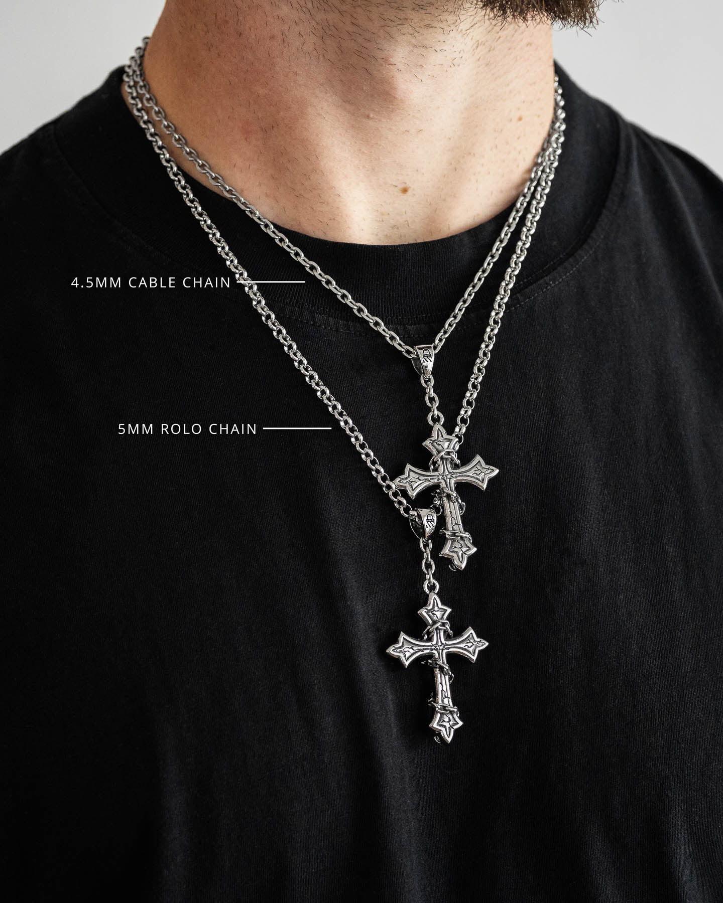 A person wearing a black t-shirt and two silver cross necklaces, each featuring "The Ruins" Pendant—one on a 4.5mm cable chain and the other on a longer 5mm rolo chain.
