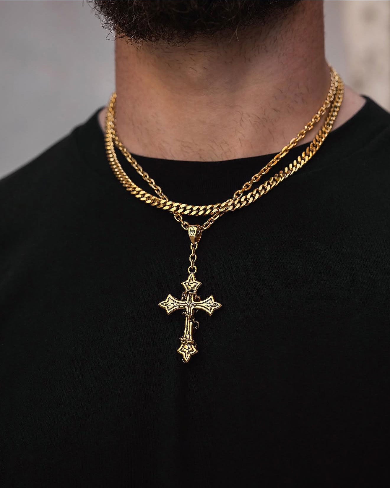 A person wearing a black shirt and layered gold chains—including Statement Collective's "The Ruins" 6mm Set (Gold) with a gothic cross pendant—is shown from the neck down, the necklace set glimmering as their beard is partly visible at the top.