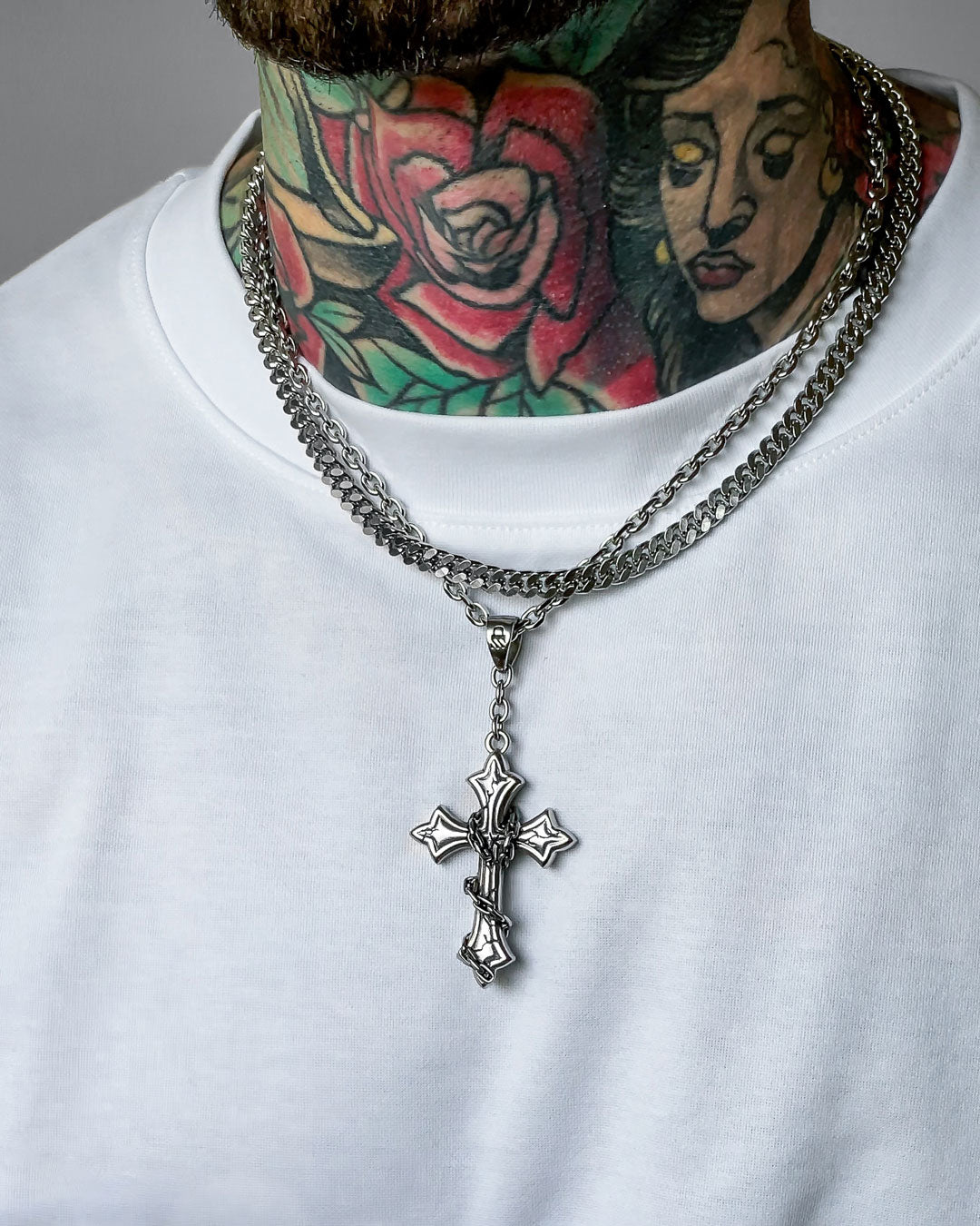 A person in a white shirt wears "The Ruins" 6mm Set—two silver chains, including a Cuban link and a large gothic cross pendant. Their neck and part of their face display colorful tattoos, like a rose and a woman's face.