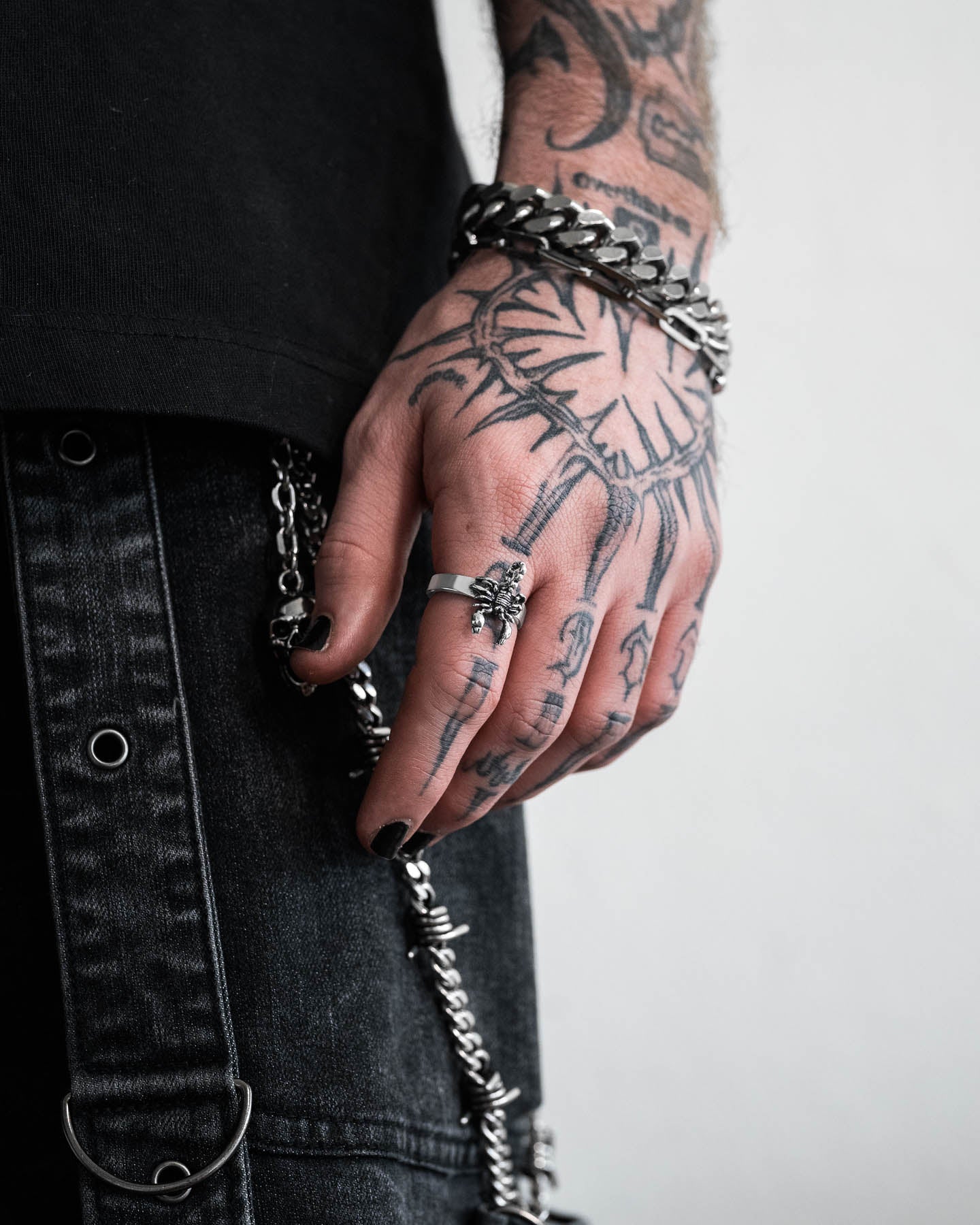 A tattooed hand with black nail polish showcases chunky silver rings, including the Scorpion Ring, and bold bracelets. The wearer’s dark clothing, metal chains, and grommets emphasize an edgy, alternative style.