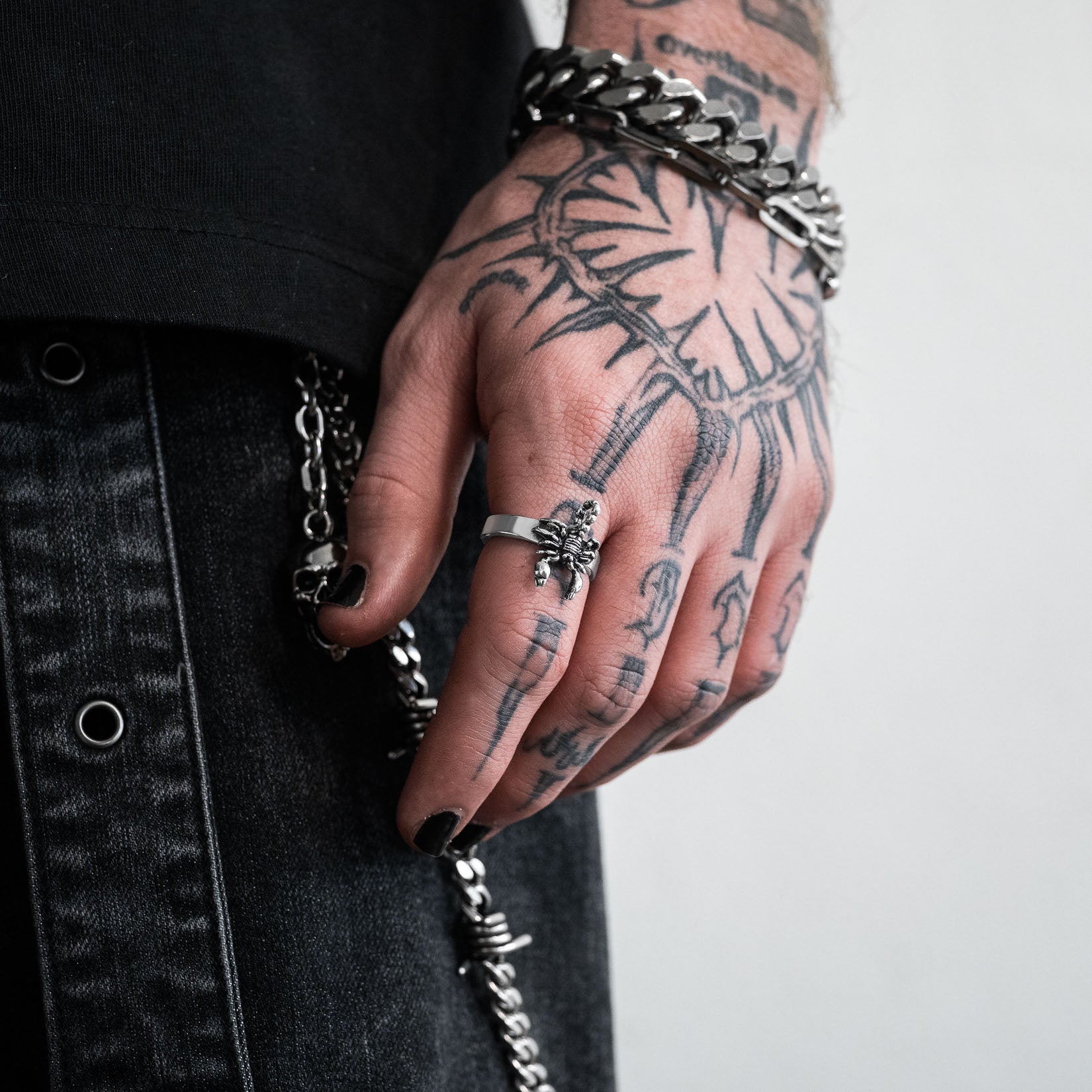 Hand with tattoos wearing a silver scorpion ring and bracelets on a plain background by statement collective