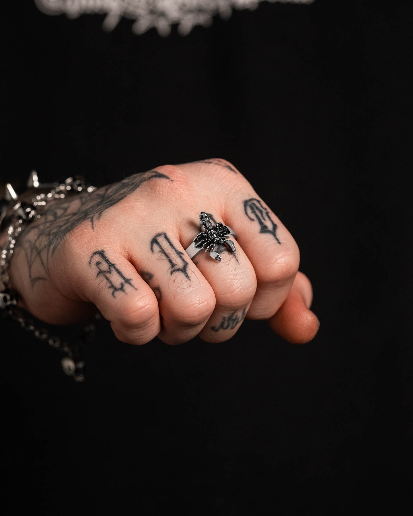 A hand with bold letter tattoos and a spiderweb on the thumb wears a detailed, dark Scorpion Ring and a stainless steel band. A spiked bracelet completes the look against a black background.