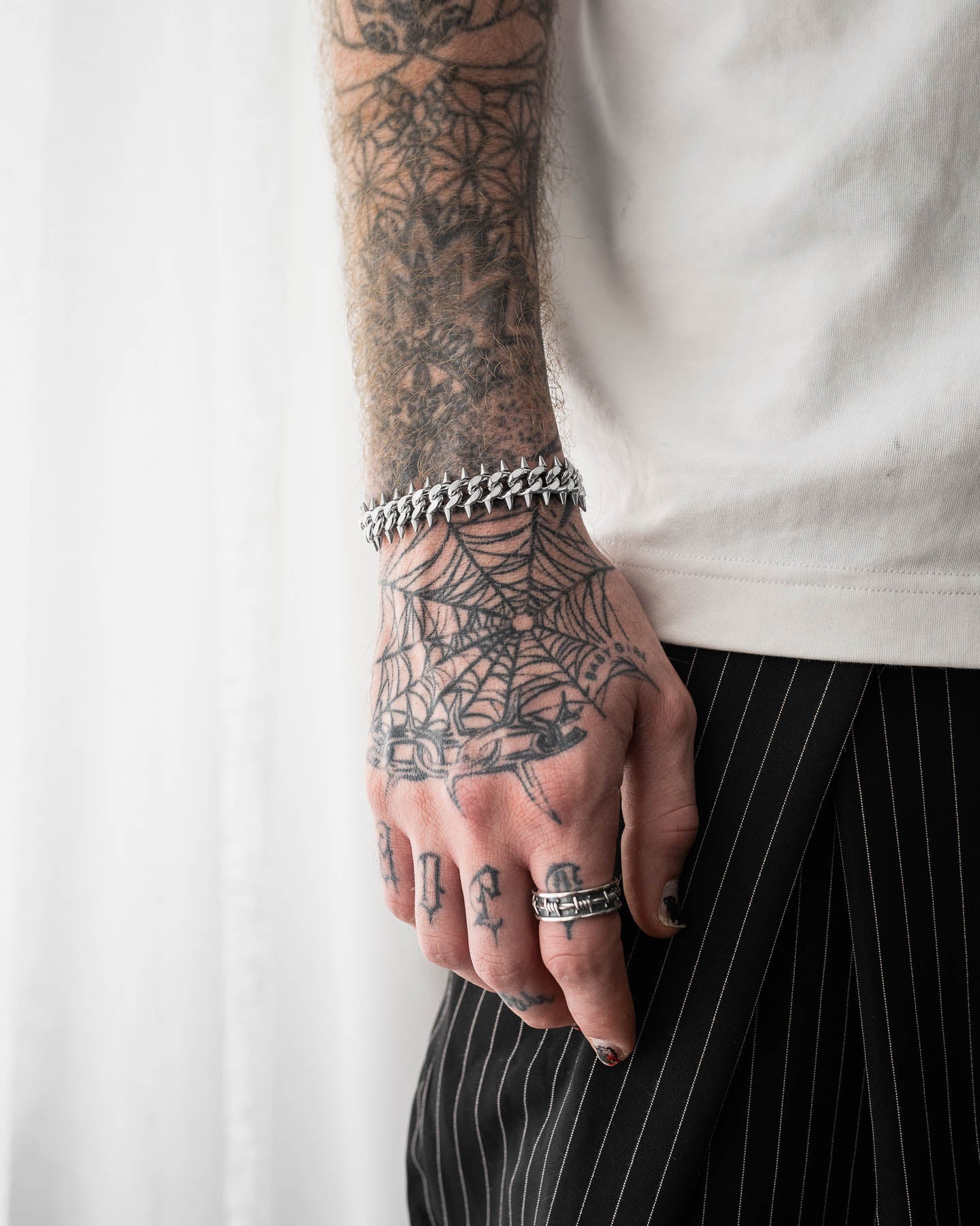 A tattooed arm and hand with rings, black nail polish, and a spiderweb design is shown wearing the "Screwed Up" Spiked Cuban Bracelet, paired with a white shirt and black pinstriped pants against a light background.