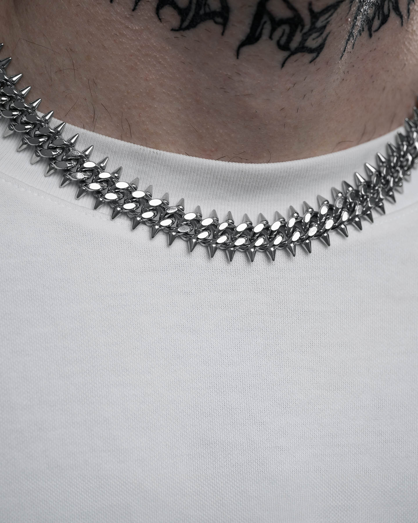 A close-up of a person wearing the "Screwed Up" Spiked Cuban Chain—a 316L stainless steel necklace with custom links—over a white shirt, black neck tattoos visible in the shot.