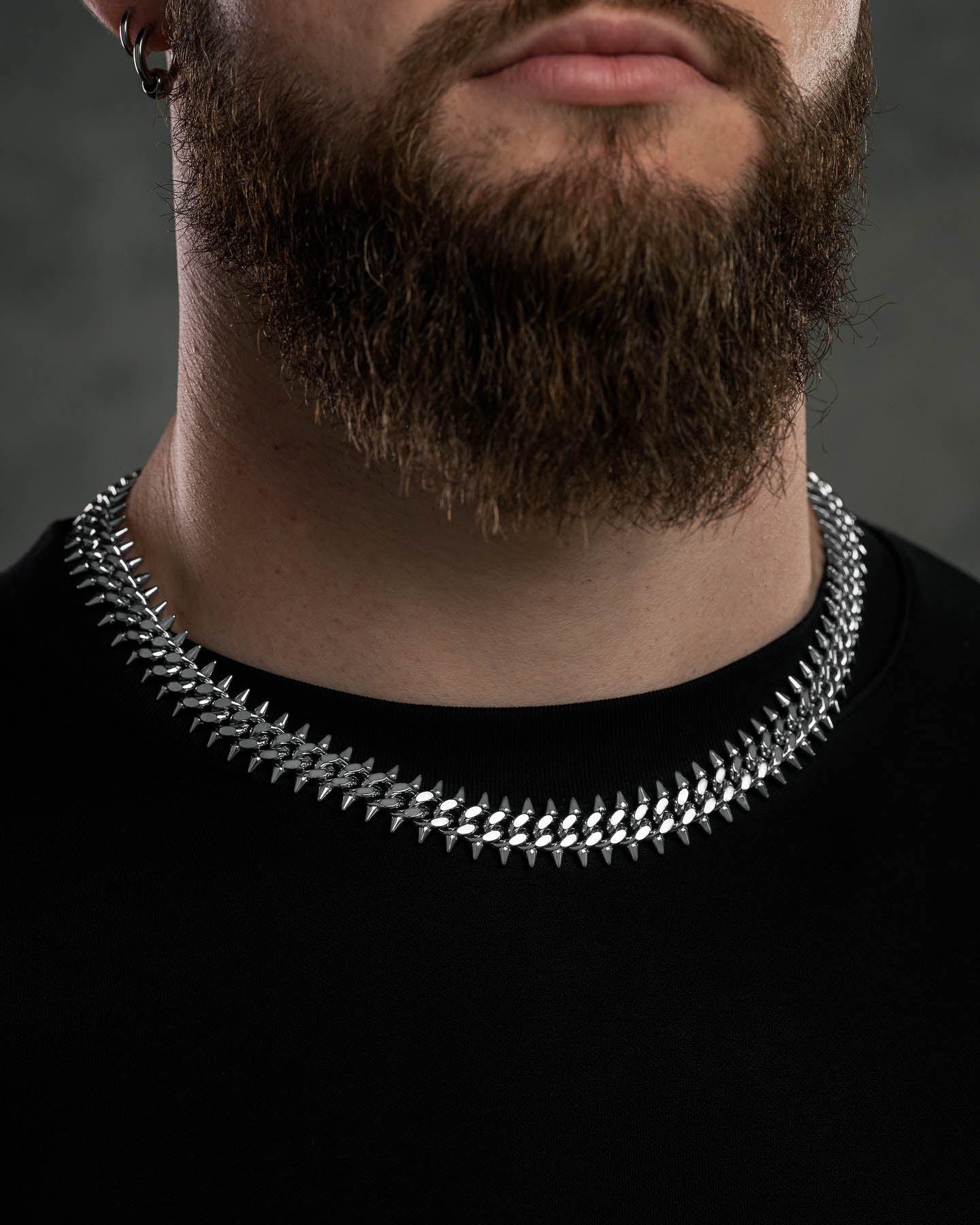 A man with a full brown beard wears a black shirt and a shiny "Screwed Up" Spiked Cuban Chain made from 316L stainless steel. The background is dark and blurred.