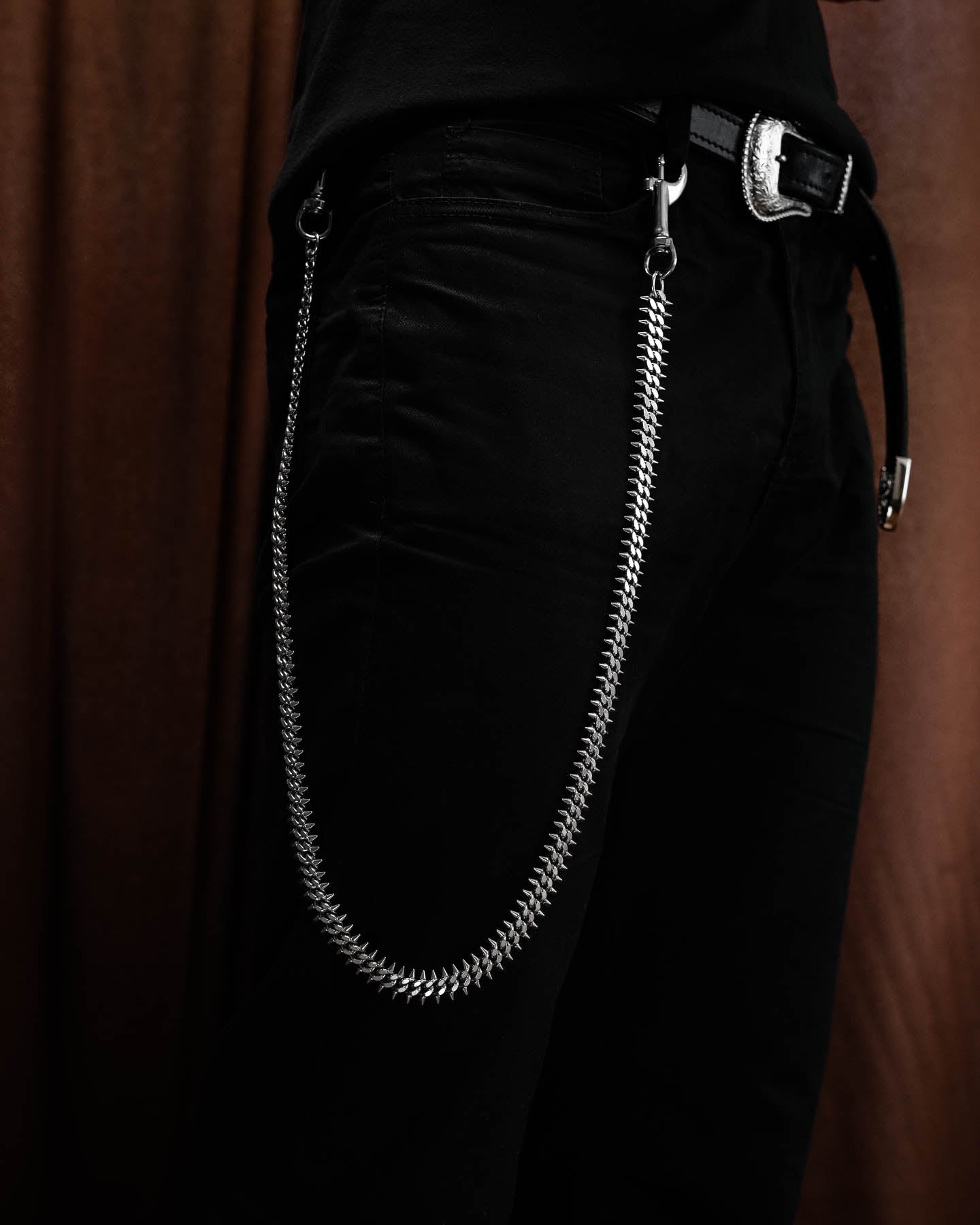 Wearing black pants and a black belt with a silver buckle, the person sports the Statement Collective "Screwed Up" 9mm Spiked Cuban Wallet Chain from their belt loop to front pocket, set against a brown curtain backdrop.