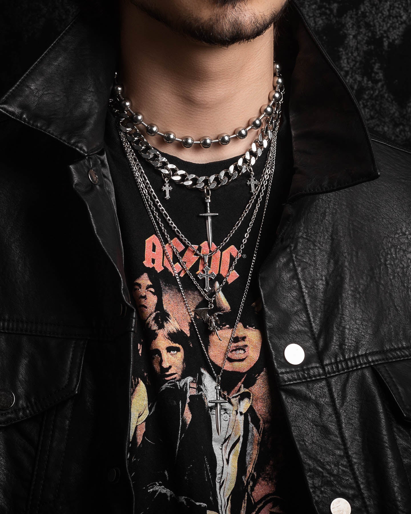 A person wearing a black leather jacket and an AC/DC tee is layered with the Sharp Objects Stack, featuring multiple silver chains and a statement cross necklace visible from neck to upper chest.