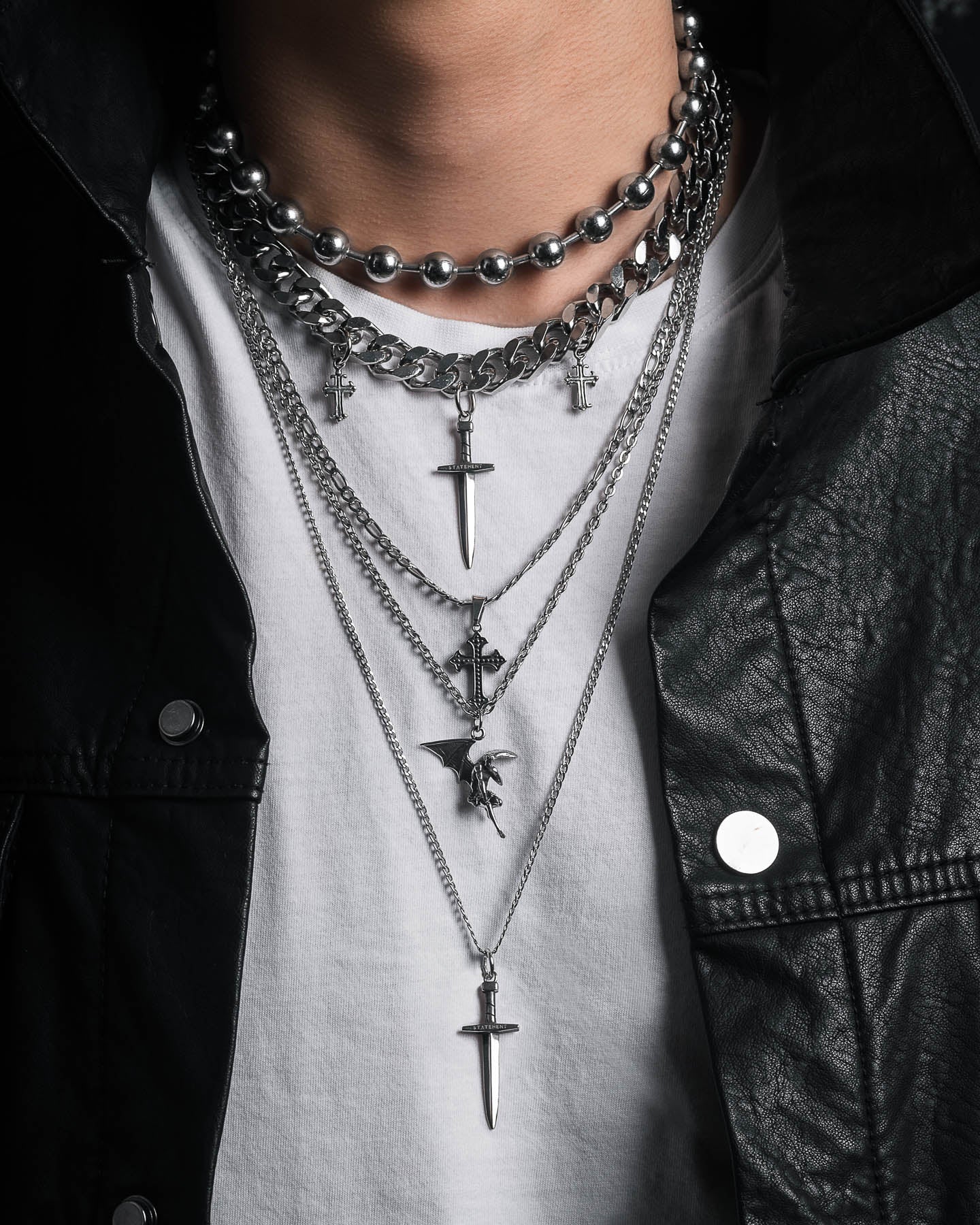 A person wearing a white t-shirt, black leather jacket, and the Sharp Objects Stack—a bold layered silver necklace set with statement pieces like crosses, daggers, and a winged figure—completes the edgy look.