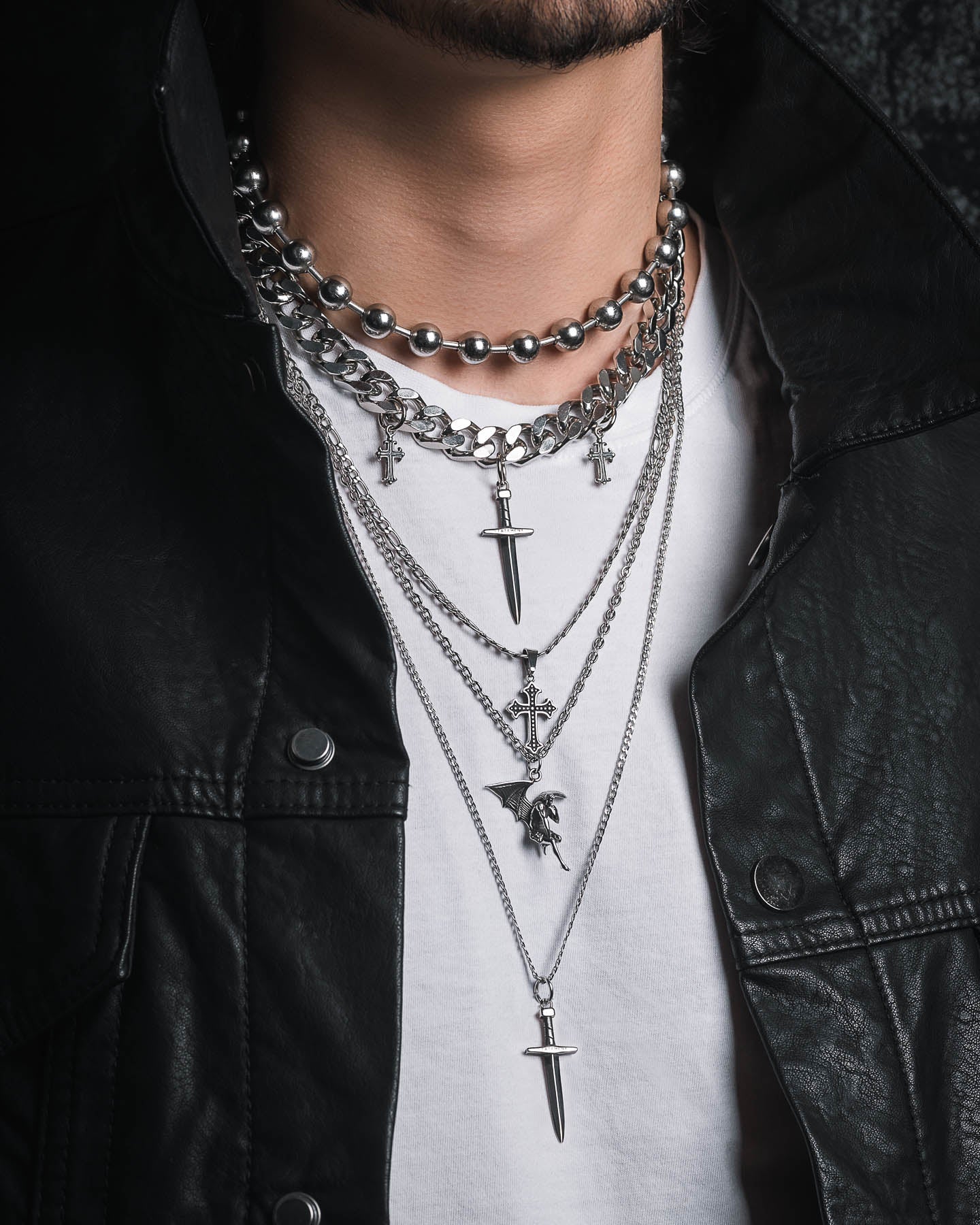 The Sharp Objects Stack features layered silver chains, beaded necklaces, a Cuban link chain, and multiple dagger and cross pendants, styled here with a black leather jacket and white shirt.