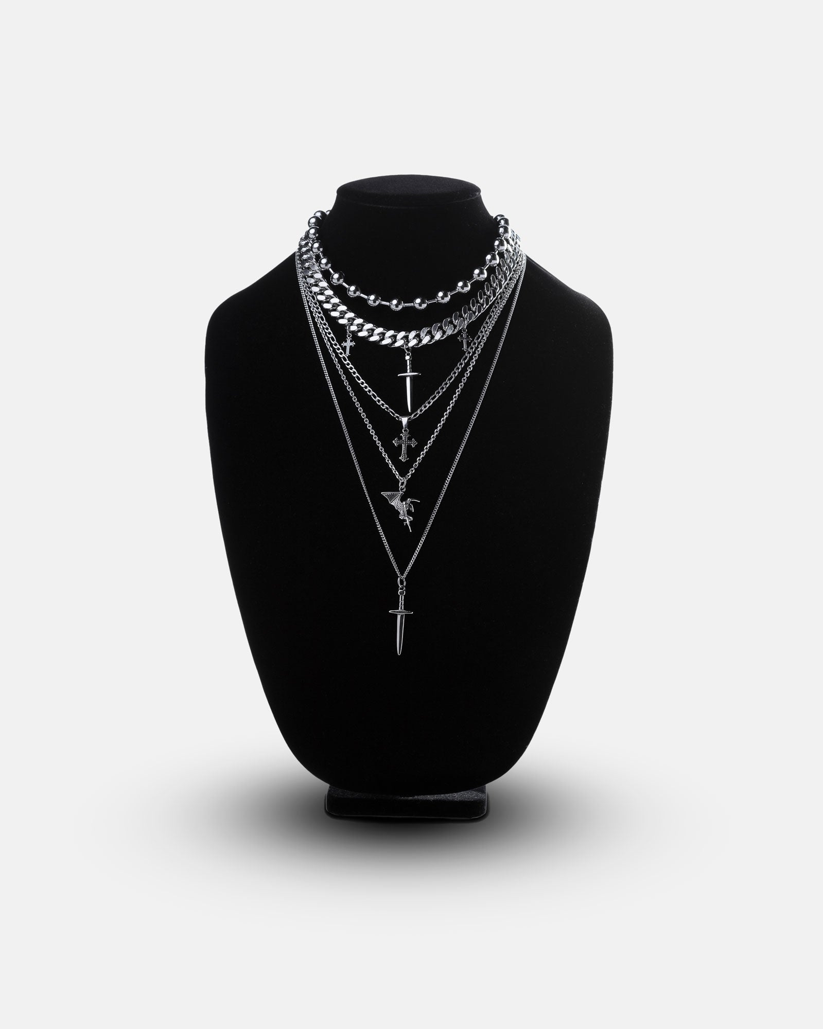 The Sharp Objects Stack, a set of four layered silver necklaces with unique pendants—a cross, sword, eagle, and a bold Cuban link chain—is displayed on a black velvet jewelry bust against a plain light gray background.