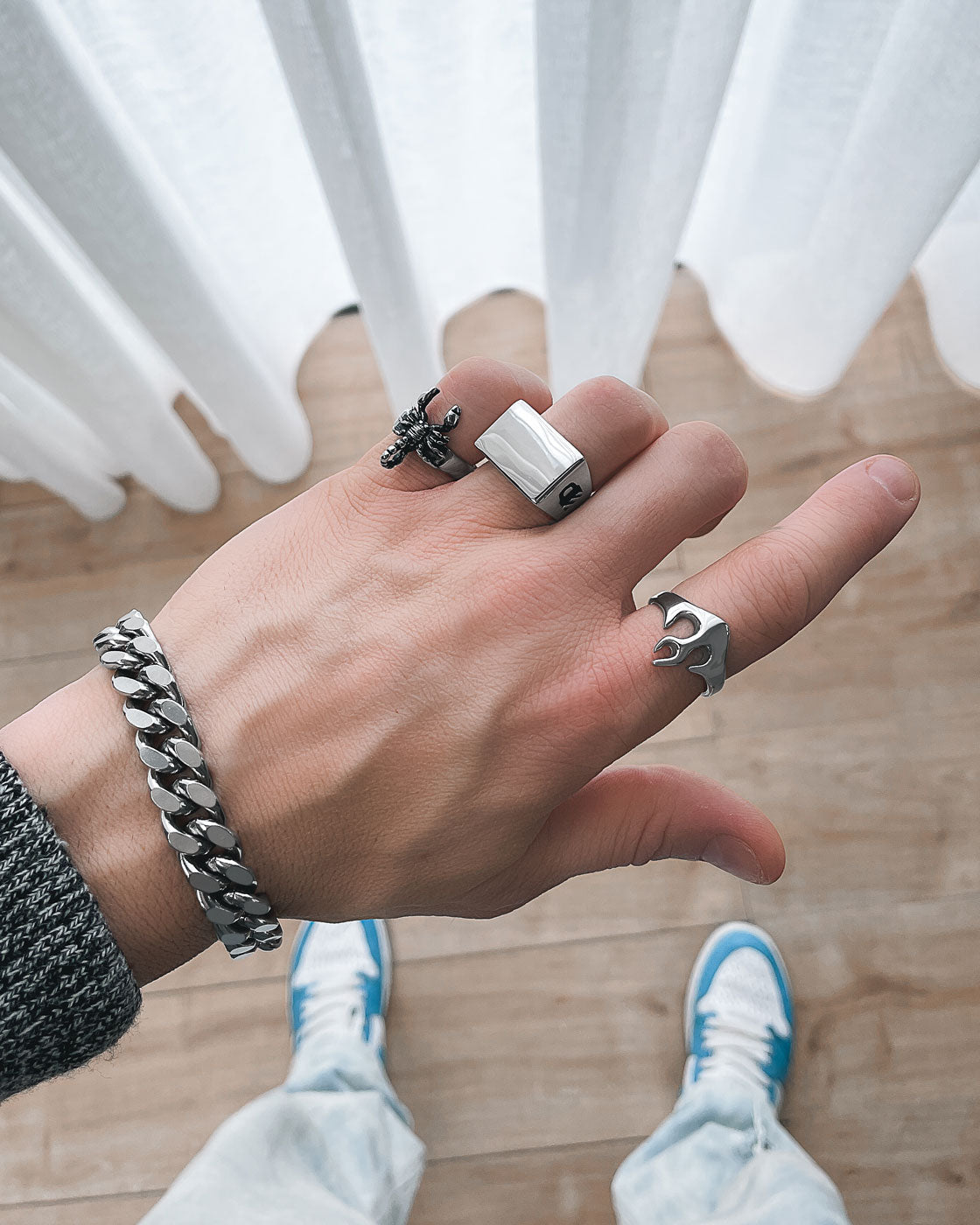 A hand wearing four silver rings, including the Statement Signet Ring, and a chunky silver bracelet is shown against a light wood floor. The person wears a dark sweater, white pants, blue and white sneakers, with white curtains in the background.