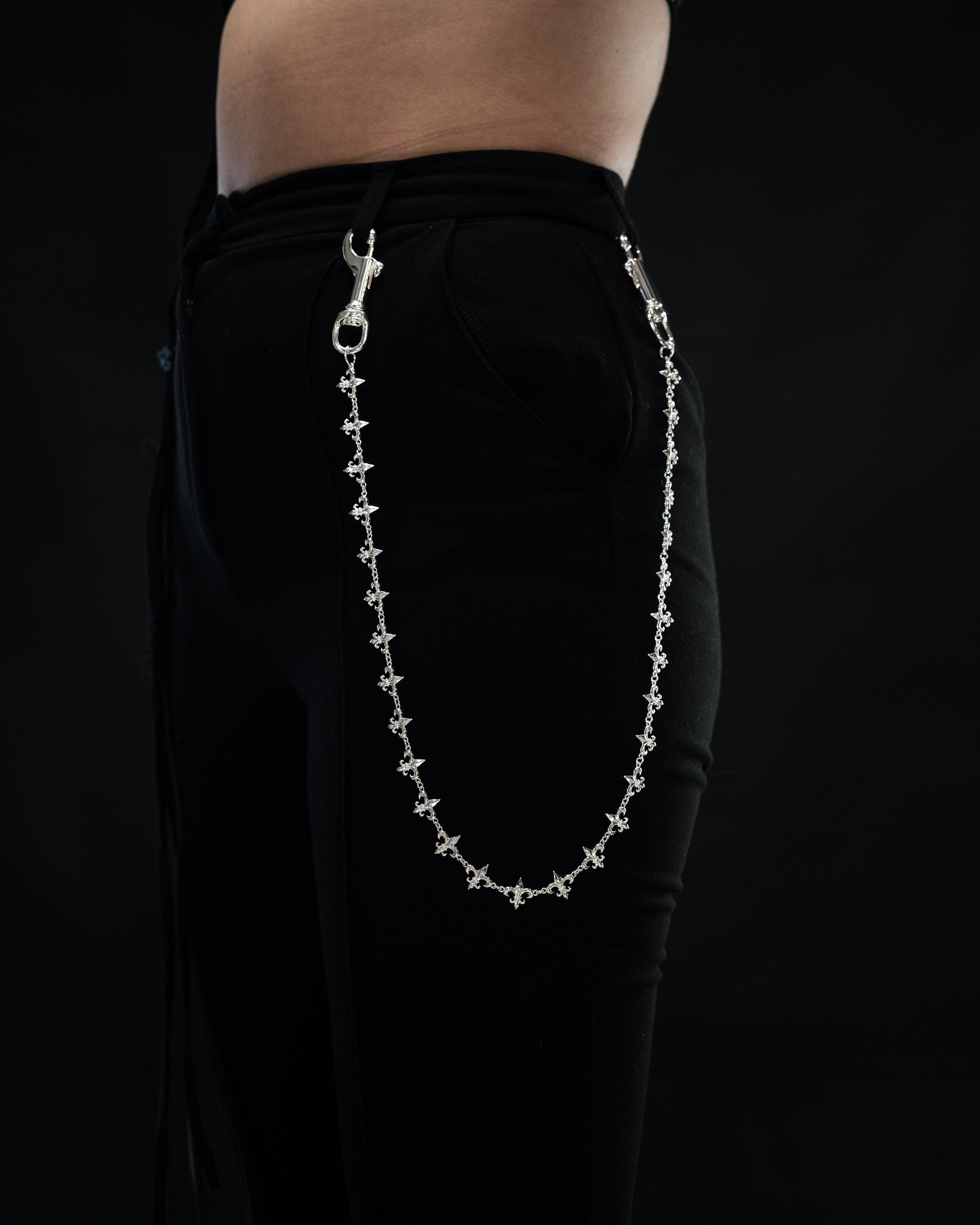 A person wearing black pants with the Statement Collective “Fleur De Lis” Wallet Chain—crafted from decorative sterling silver—attached from the front pocket to a belt loop, set against a dark background.