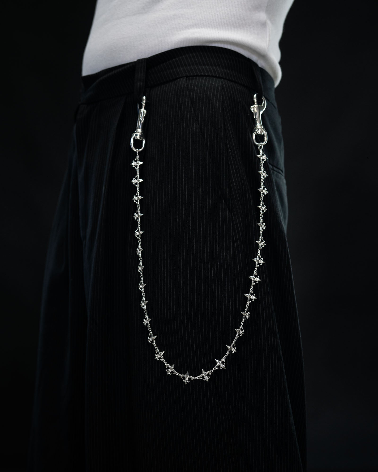 A person wearing black pinstripe pants with the “Fleur De Lis” Wallet Chain by Statement Collective, crafted from decorative sterling silver and attached at the waistband, standing against a dark background.