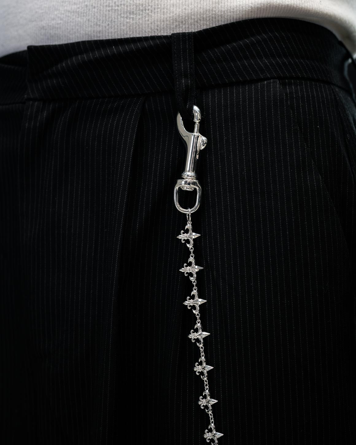 A close-up of a person wearing black pinstripe pants with the ornate Statement Collective “Fleur De Lis” Wallet Chain hanging from a belt loop, and a white shirt tucked in at the waist.