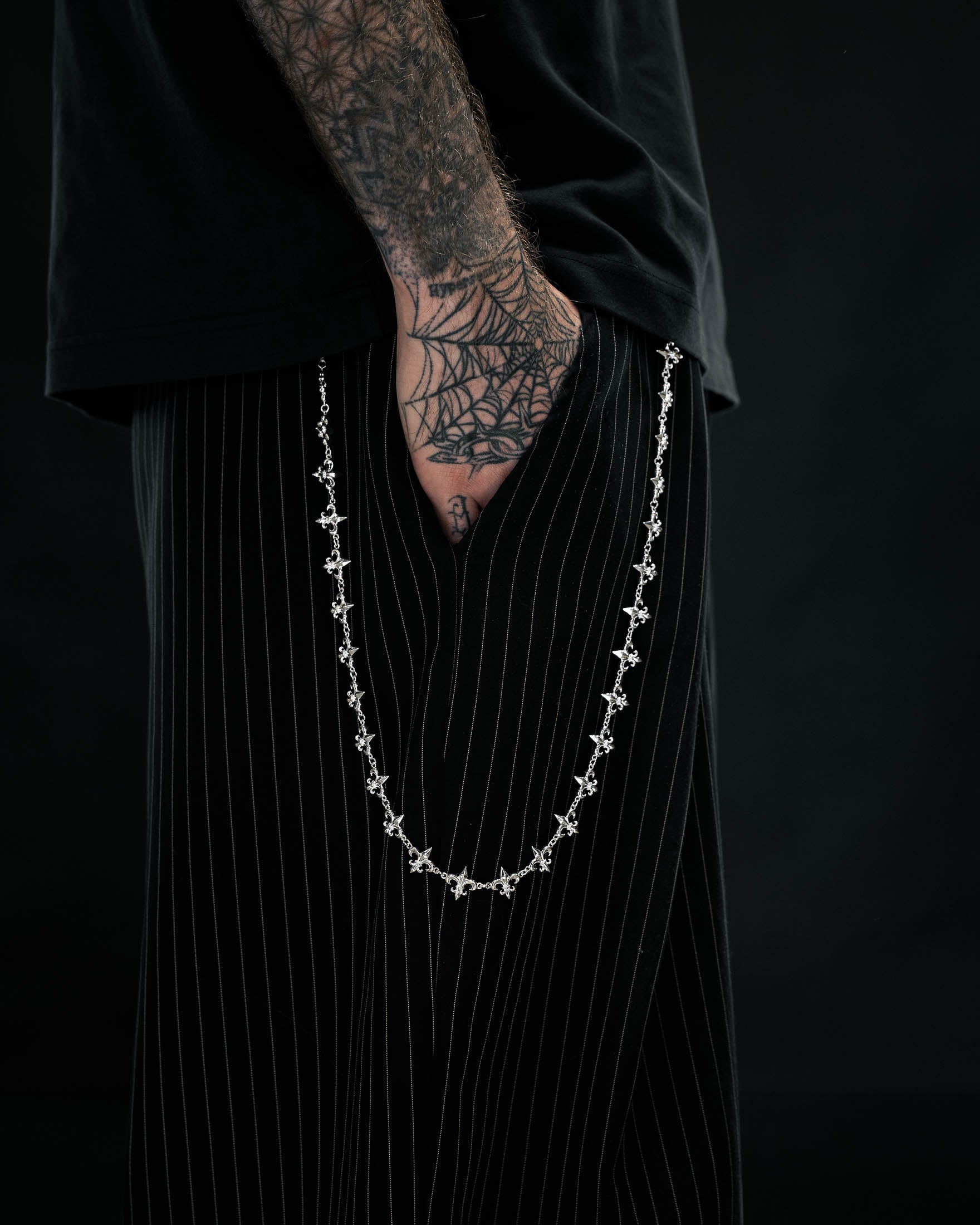 A person with a spiderweb tattoo on their hand wears black pinstripe pants, a black shirt, and the ornate “Fleur De Lis” Wallet Chain by Statement Collective hanging from their pocket, set against a dark background.