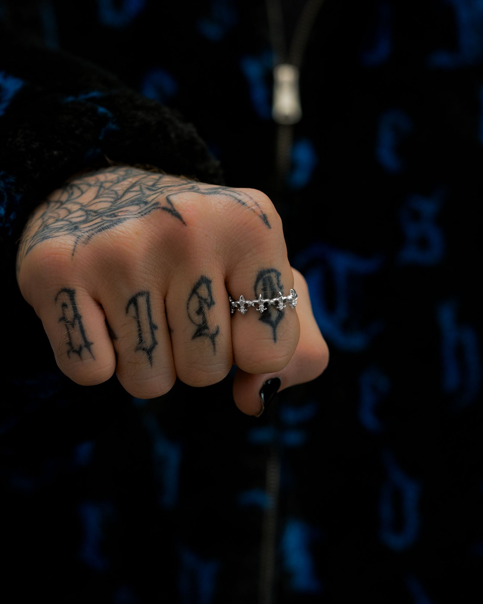 A close-up of a tattooed hand with a spiderweb design and bold letters on the fingers, adorned with Statement Collective’s striking “Fleur De Lis” silver ring alongside a sleek stainless steel band. The background features dark fabric with blue patterns.