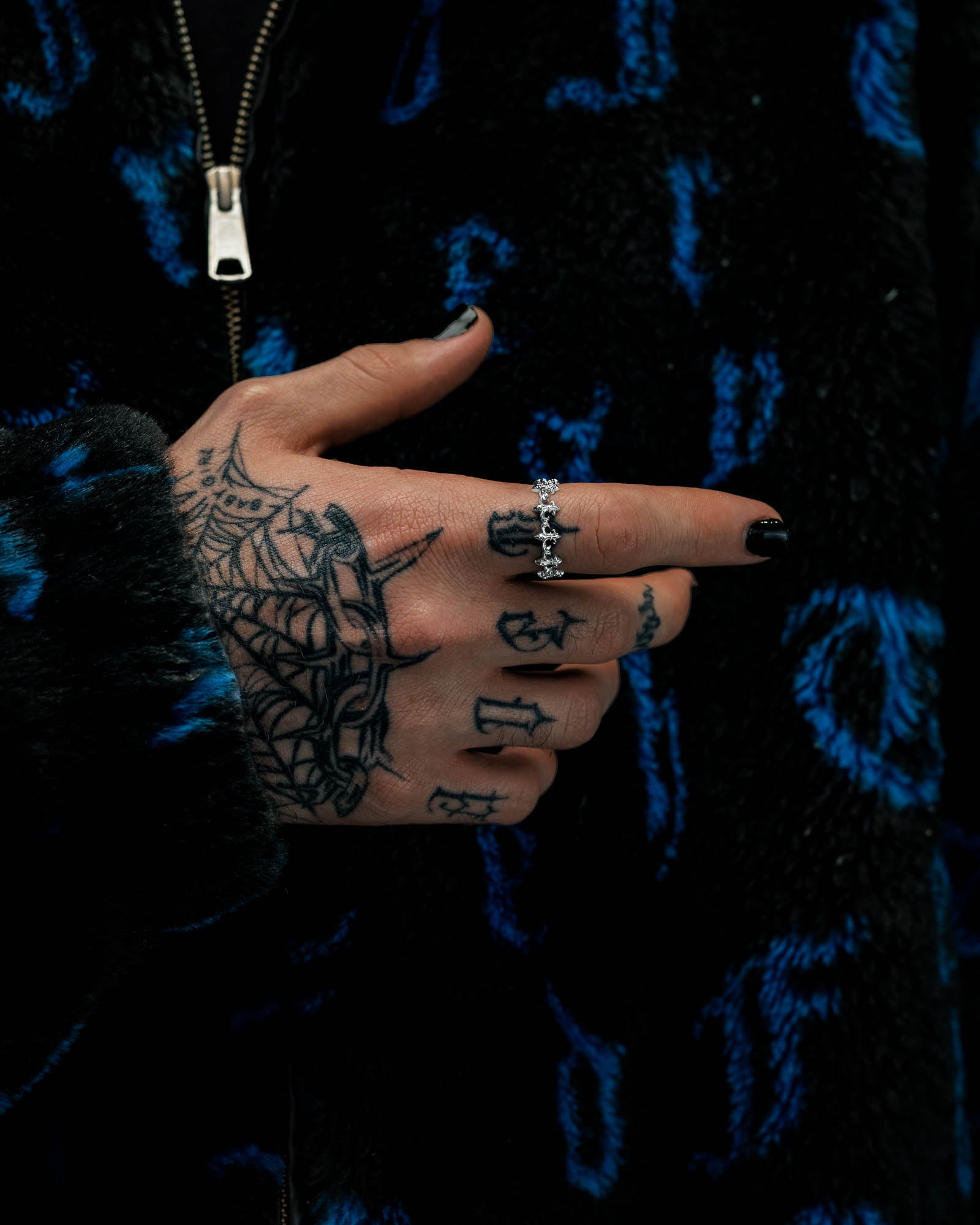 A hand adorned with spider web tattoos and black nail polish showcases the “Fleur De Lis” Ring by Statement Collective—a striking stainless steel piece encrusted with shimmering diamonds. The person wears a black jacket with blue patterns, featuring a partially visible zipper.