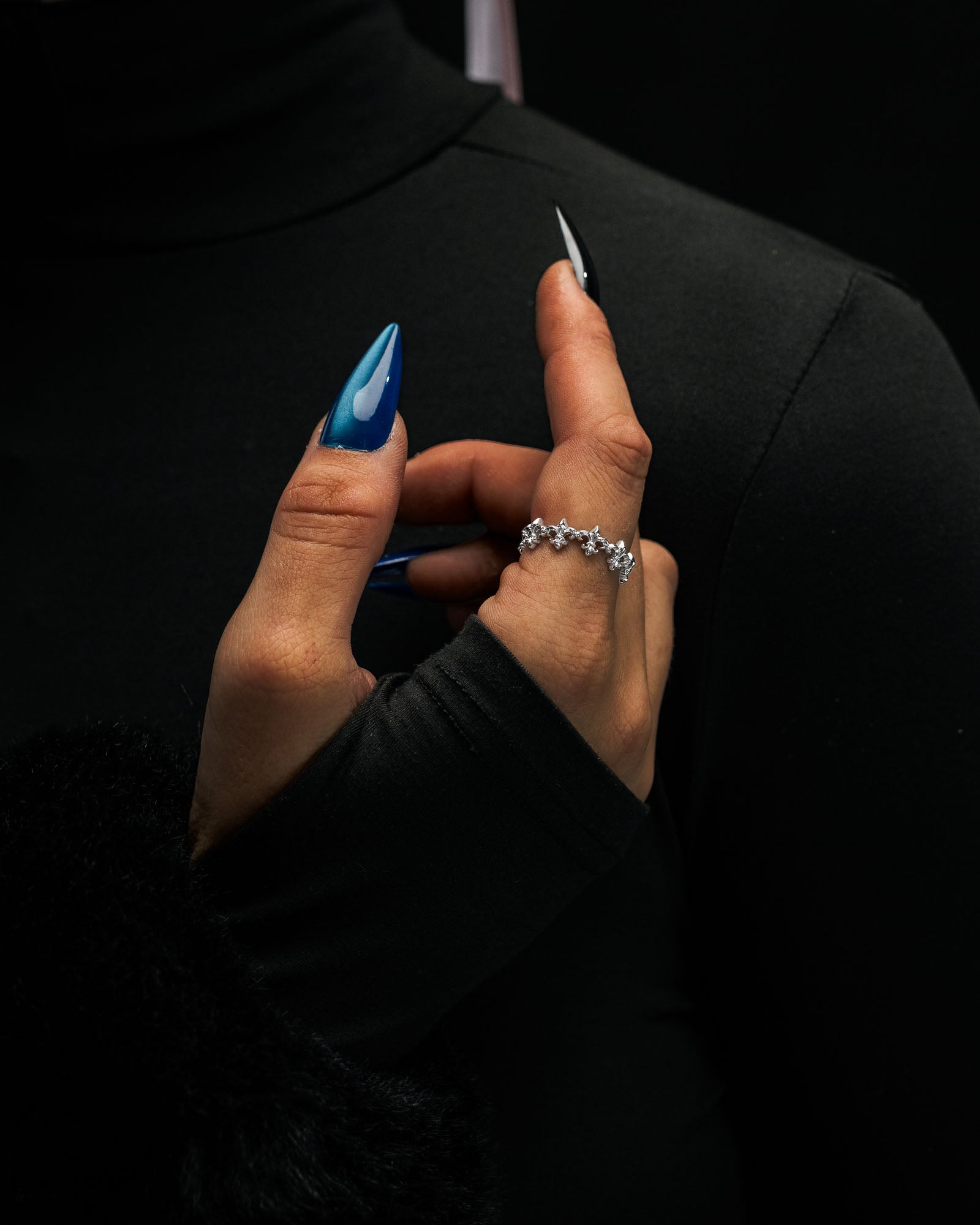 A hand with long blue nails showcases the “Fleur De Lis” ring by Statement Collective, a delicate stackable piece adorned with small gemstones, posed against a black turtleneck and sleeve background.