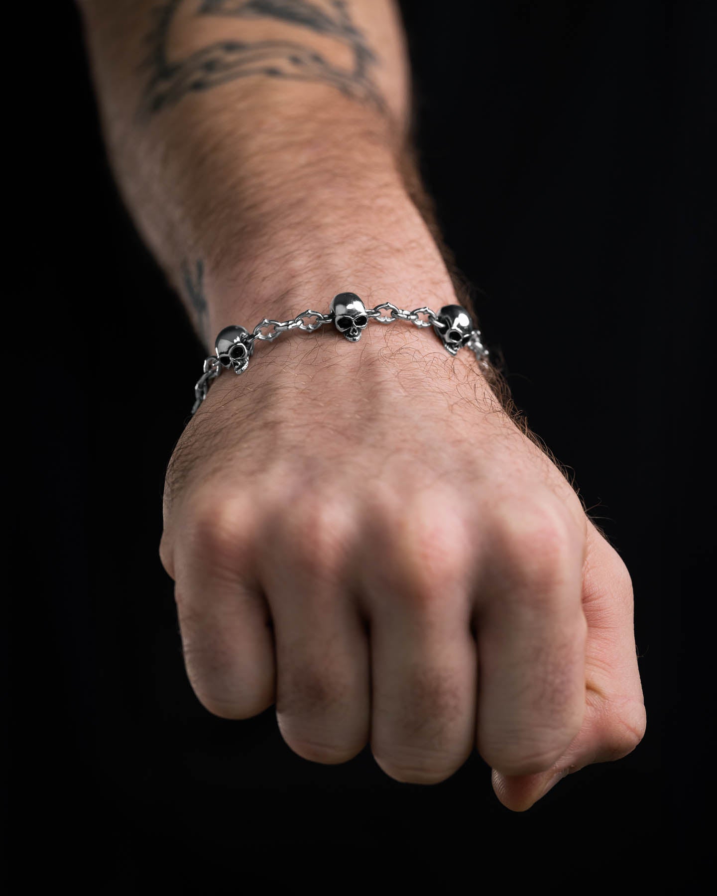 A close-up of a fist wearing the Skull Charm "Cathedral" Spiked Link Bracelet. Small skull charms and spikes stand out, with a partially visible forearm tattoo against a black background, highlighting the bracelet’s bold alternative style.