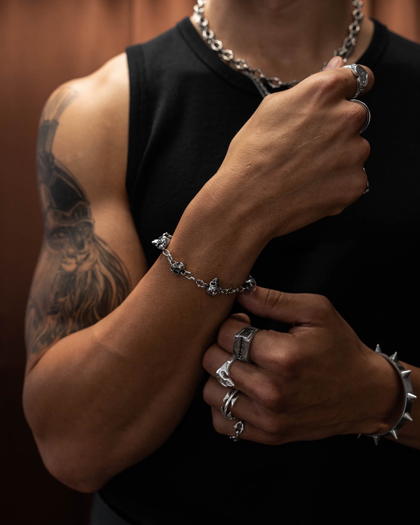 A muscular person in a black sleeveless shirt displays alternative style jewelry, including rings, a "Cathedral" Spiked Link Skull Charm Bracelet, and a chain necklace. Their left arm features a large tattoo against a softly blurred background.