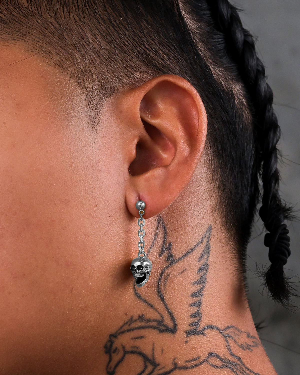 A silver skull pendant from a pair of Statement Collective Skull Drop Earrings hangs from a chain earring in an earlobe with a rock-themed design.
