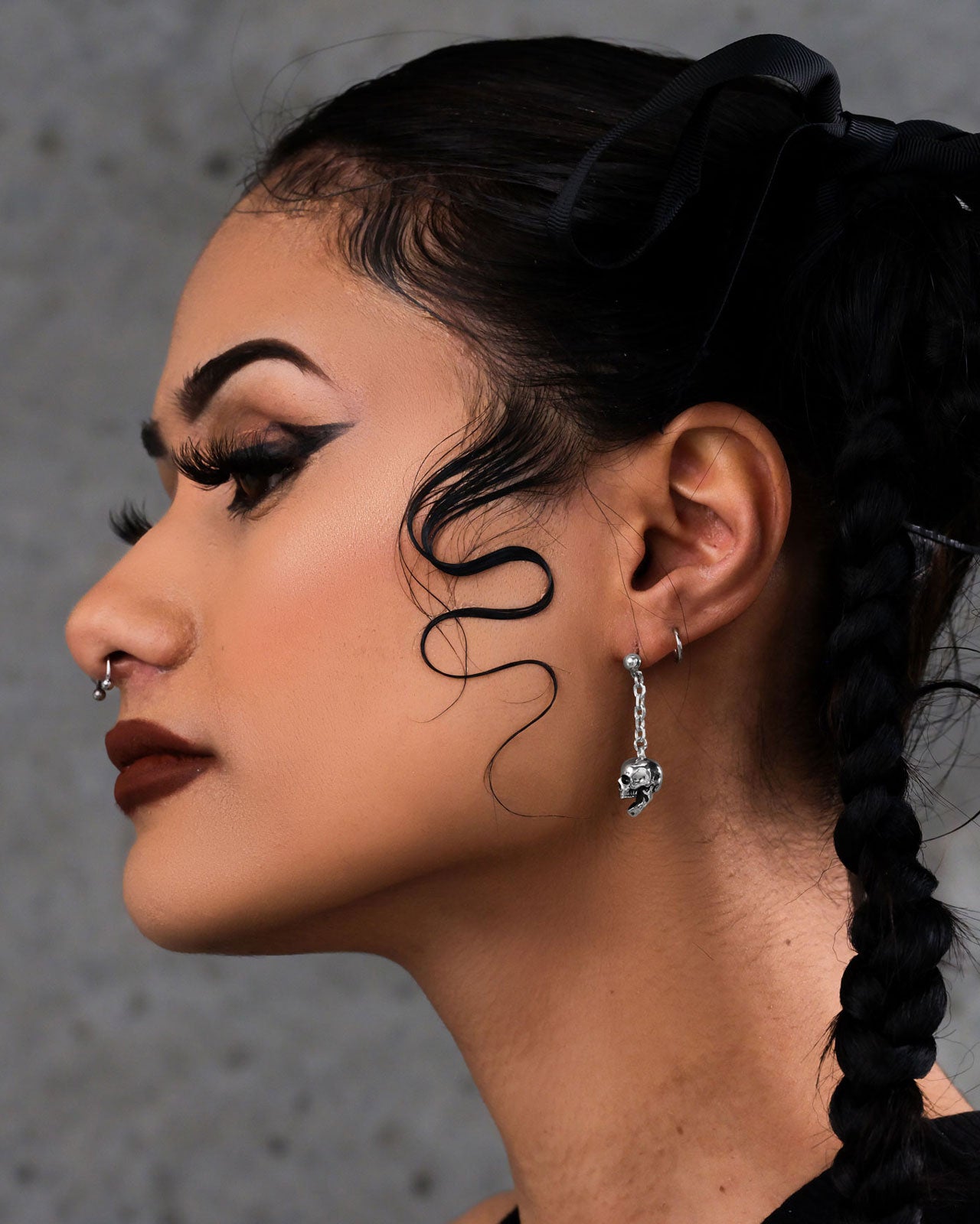 A woman with bold makeup, winged eyeliner, dark lipstick, braids, and a nose ring is shown in profile wearing striking Skull Drop Earrings. Her baby hairs are styled and the background is gray and textured.