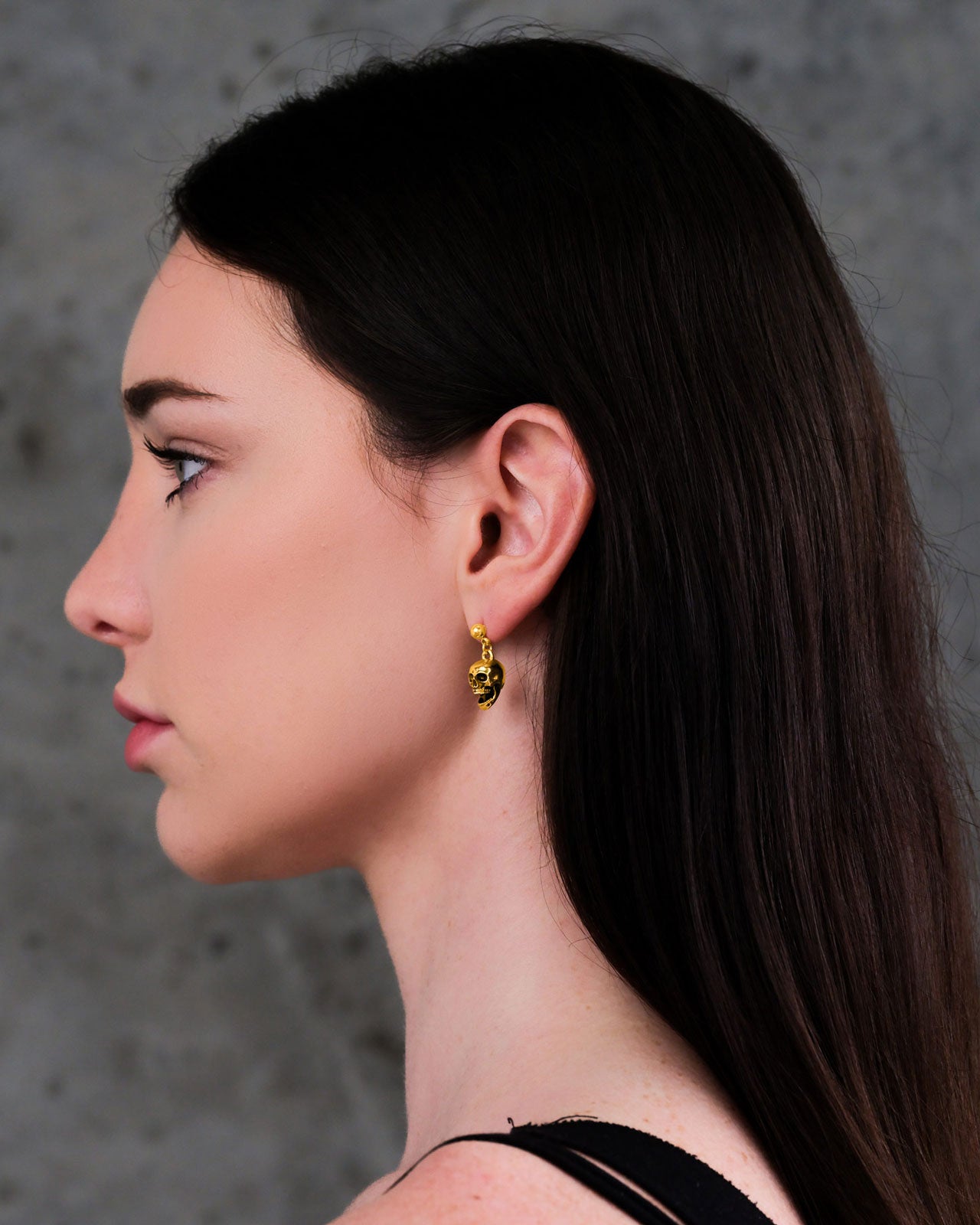 A woman with long dark hair wears Skull Earrings (Gold), gazing calmly ahead against a gray textured background and showcasing her subtle punk jewelry style.