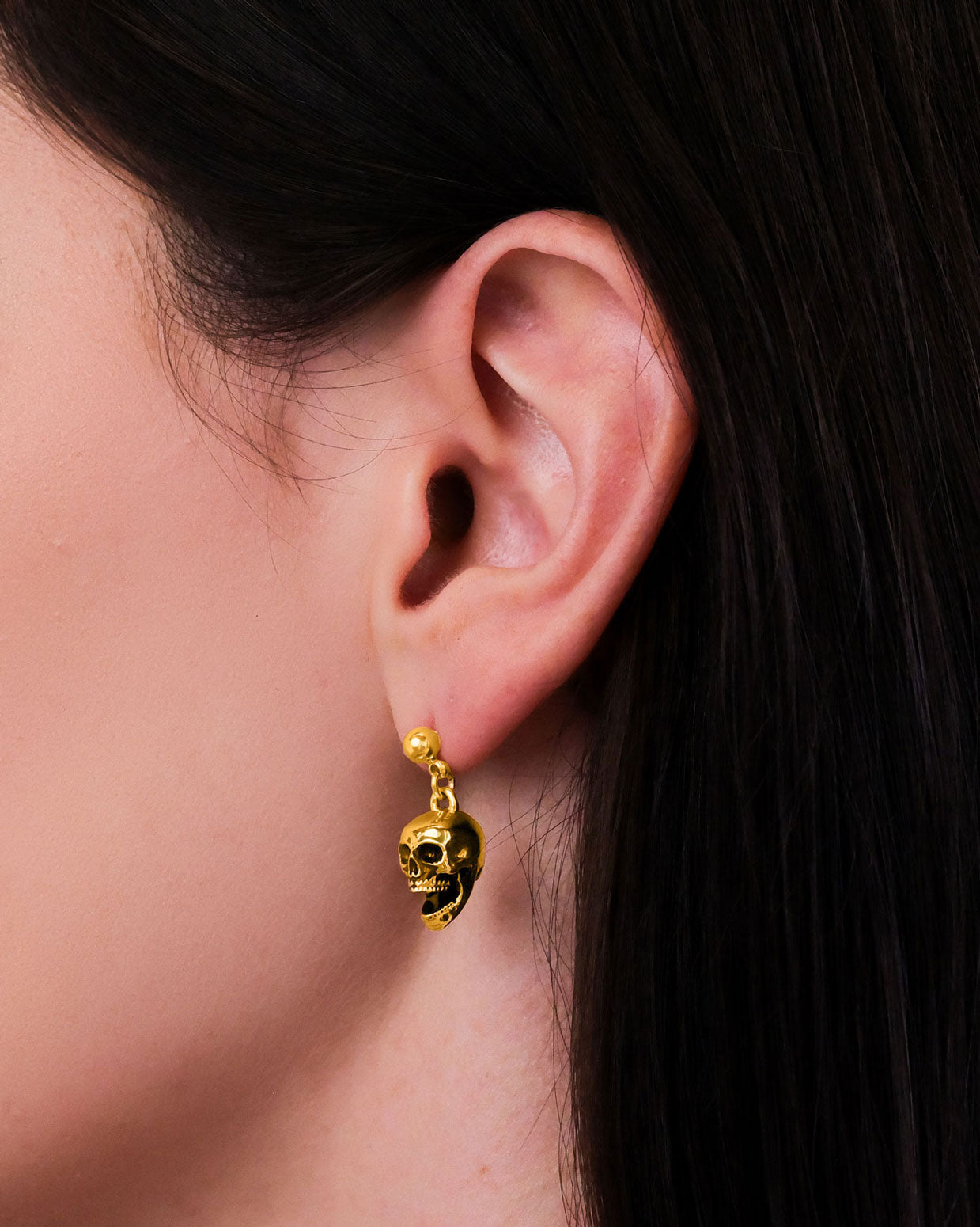 Close-up of a person's ear with straight dark brown hair, wearing Skull Earrings (Gold)—18k gold-plated accessories that make a bold punk statement.