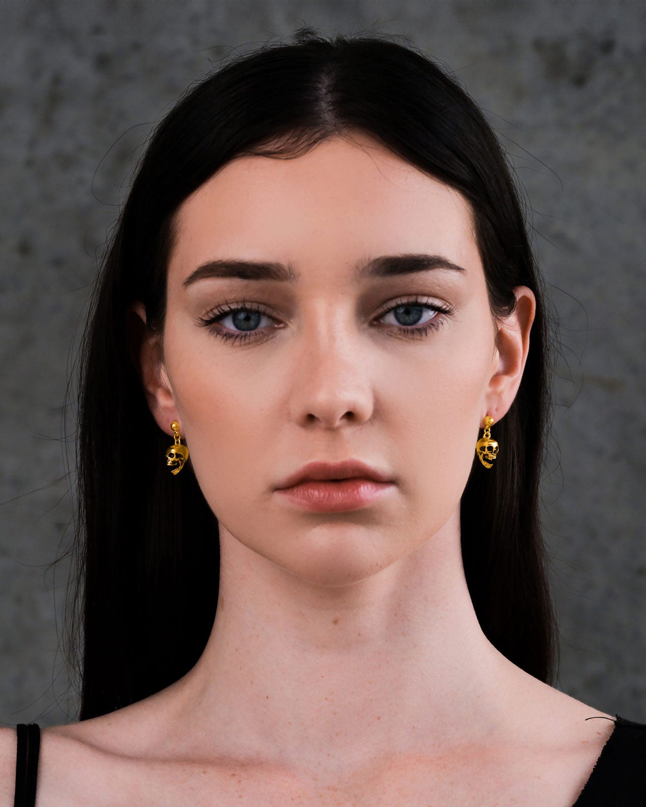 A woman with straight dark hair and light skin faces the camera, wearing Skull Earrings (Gold) against a gray textured background.