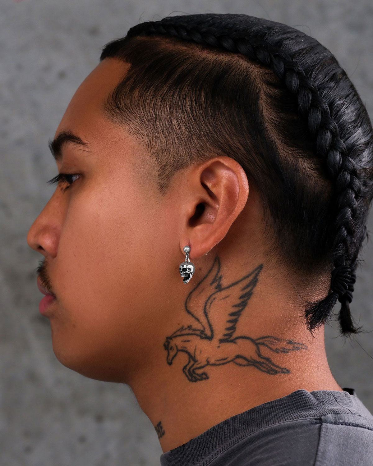 A person with braided hair, a mustache, and a Pegasus neck tattoo is shown in profile wearing Skull Earrings made from hypoallergenic stainless steel. They have on a gray shirt against a plain gray background.