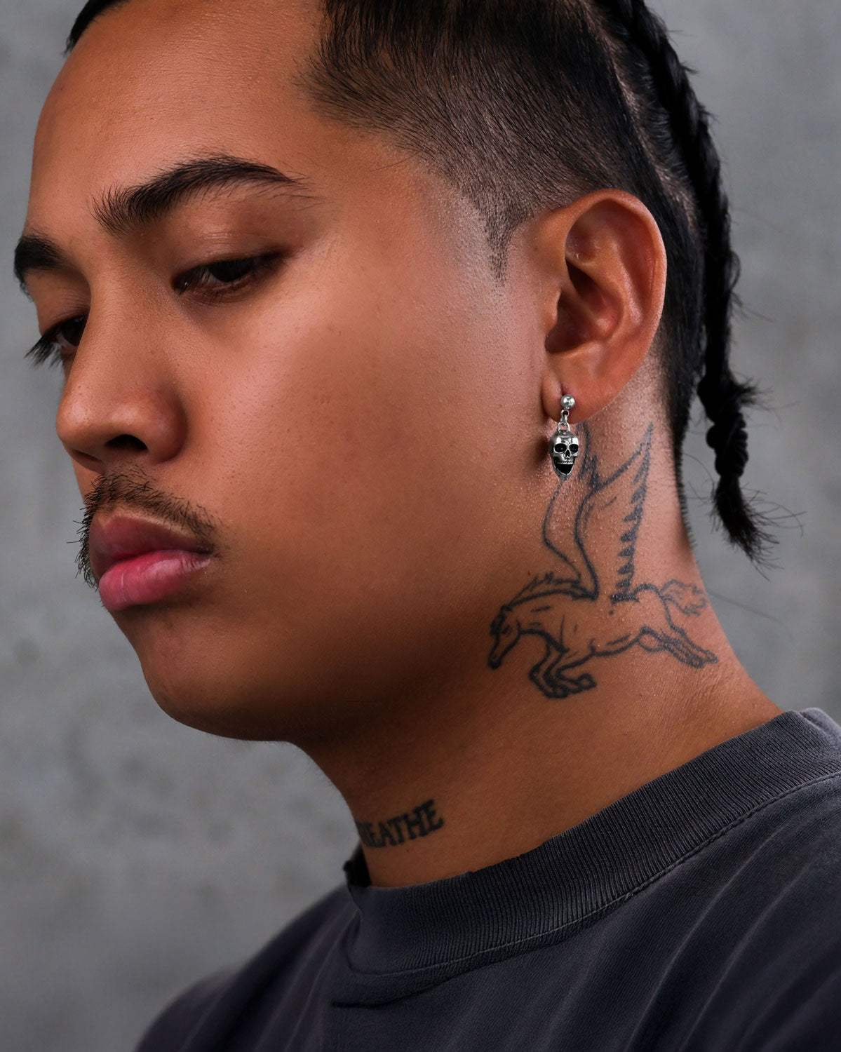 A person with braided hair and a mustache wears a dark shirt, gazes downward, and has a pegasus tattoo on their neck and a word tattoo on their throat. They accessorize with Skull Earrings. The background is gray.