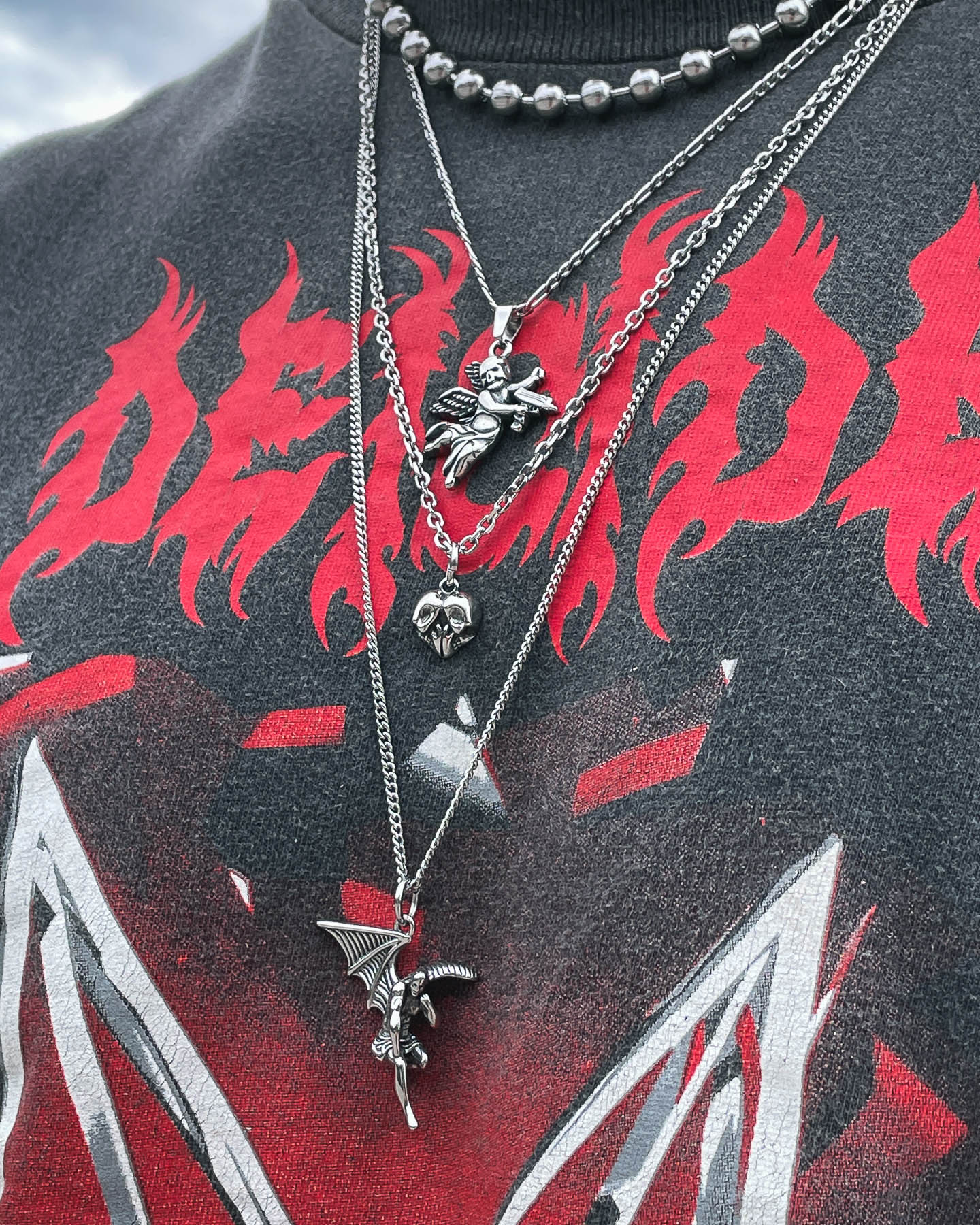 Close-up of a person wearing the Skull Heart Pendant—one of four layered silver necklaces—over a black shirt with bold red flame-like designs and text.