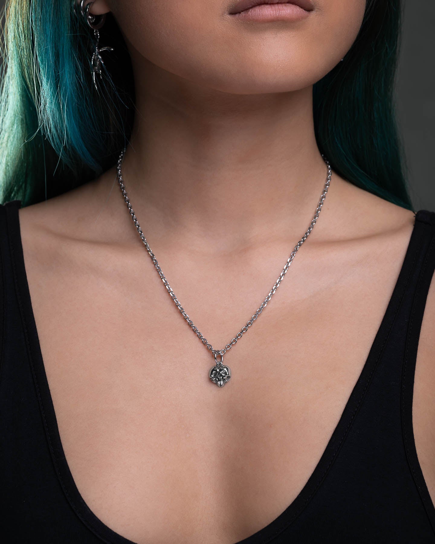 A woman with teal hair models the Skull Heart Pendant—a stainless steel chain with a small ornate skull heart—paired with a black tank top. Only her lower face, neck, and upper chest are visible against a dark background.