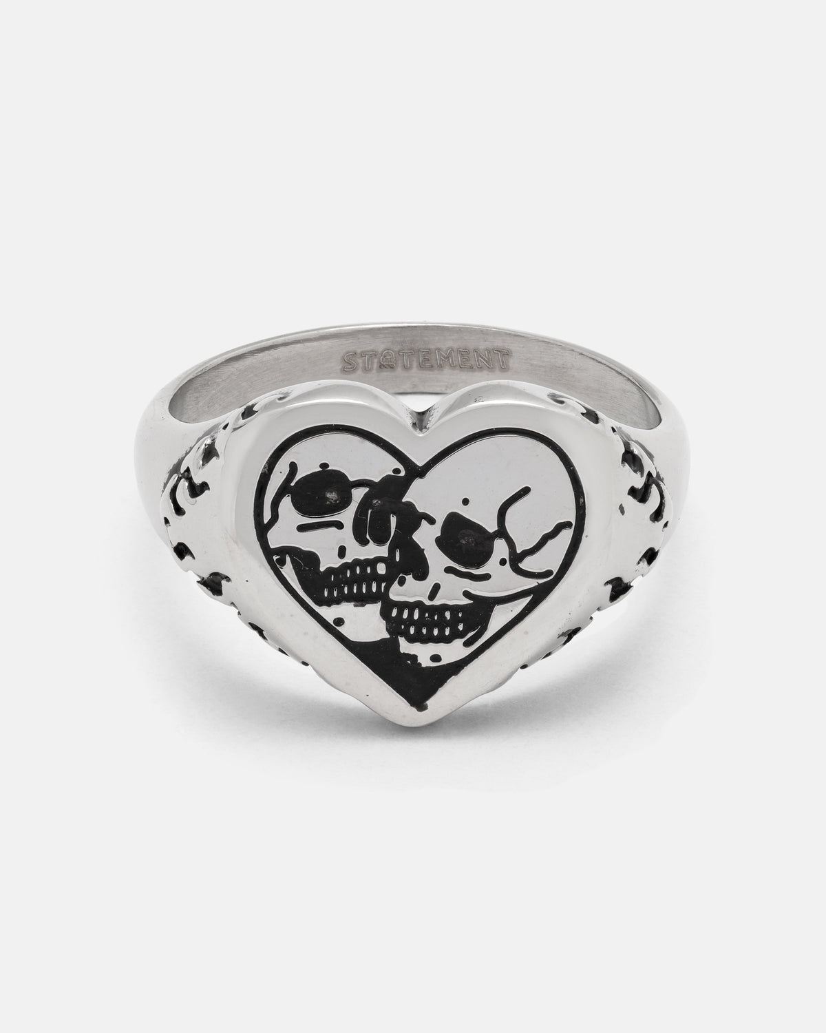 The STATEMENT Skull Heart Ring is a silver heart-shaped ring showcasing two black skulls facing each other engraved on the front, accented with subtle decorative markings along the sides. This bold piece of gothic jewelry is perfect for those who embrace unique and unconventional style.