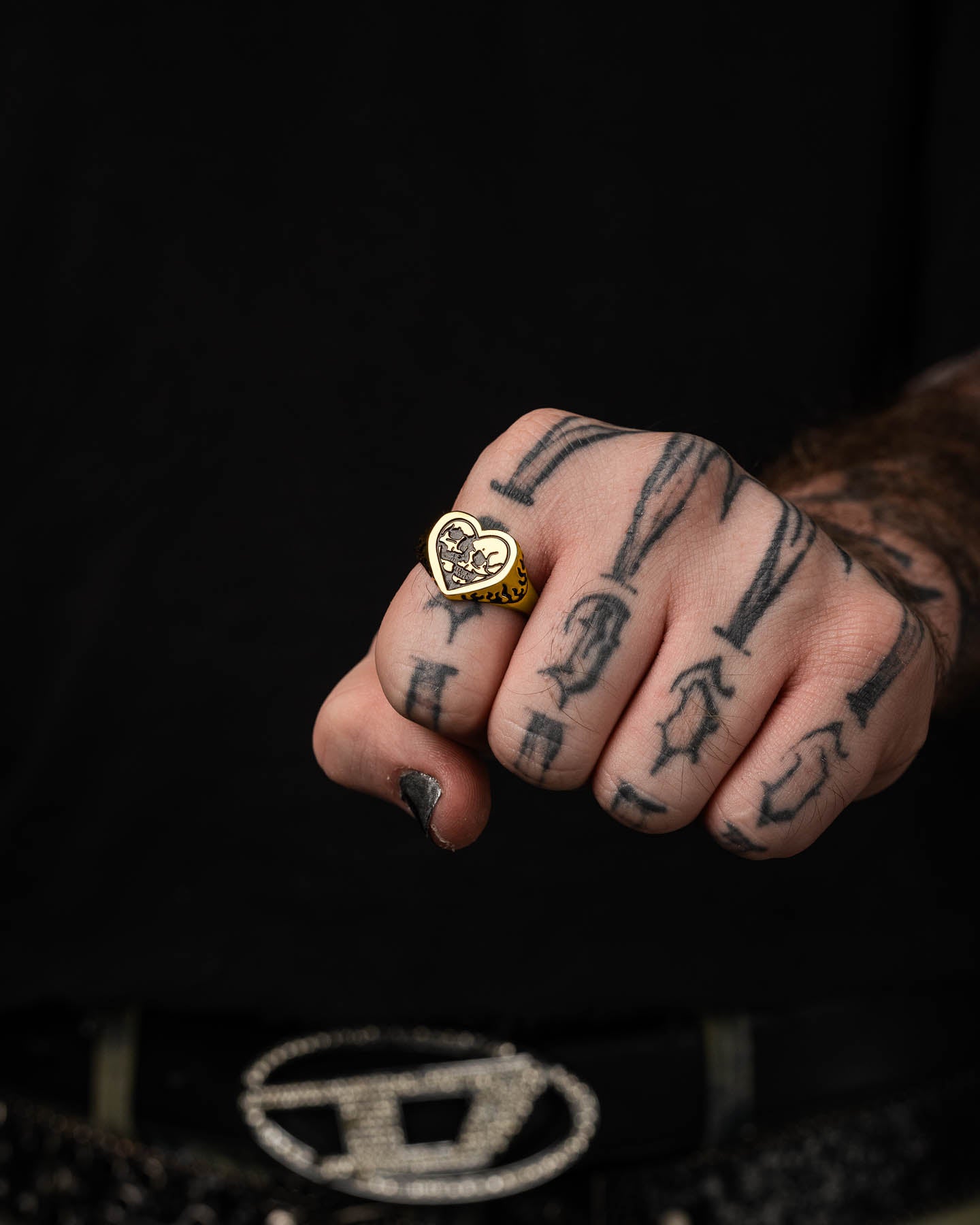 A hand with black-painted nails and tattoos holds the Skull Heart Ring (Gold). The person, dressed in a black shirt and bold belt, highlights their unique gothic jewelry style with this striking 18k gold plated piece.