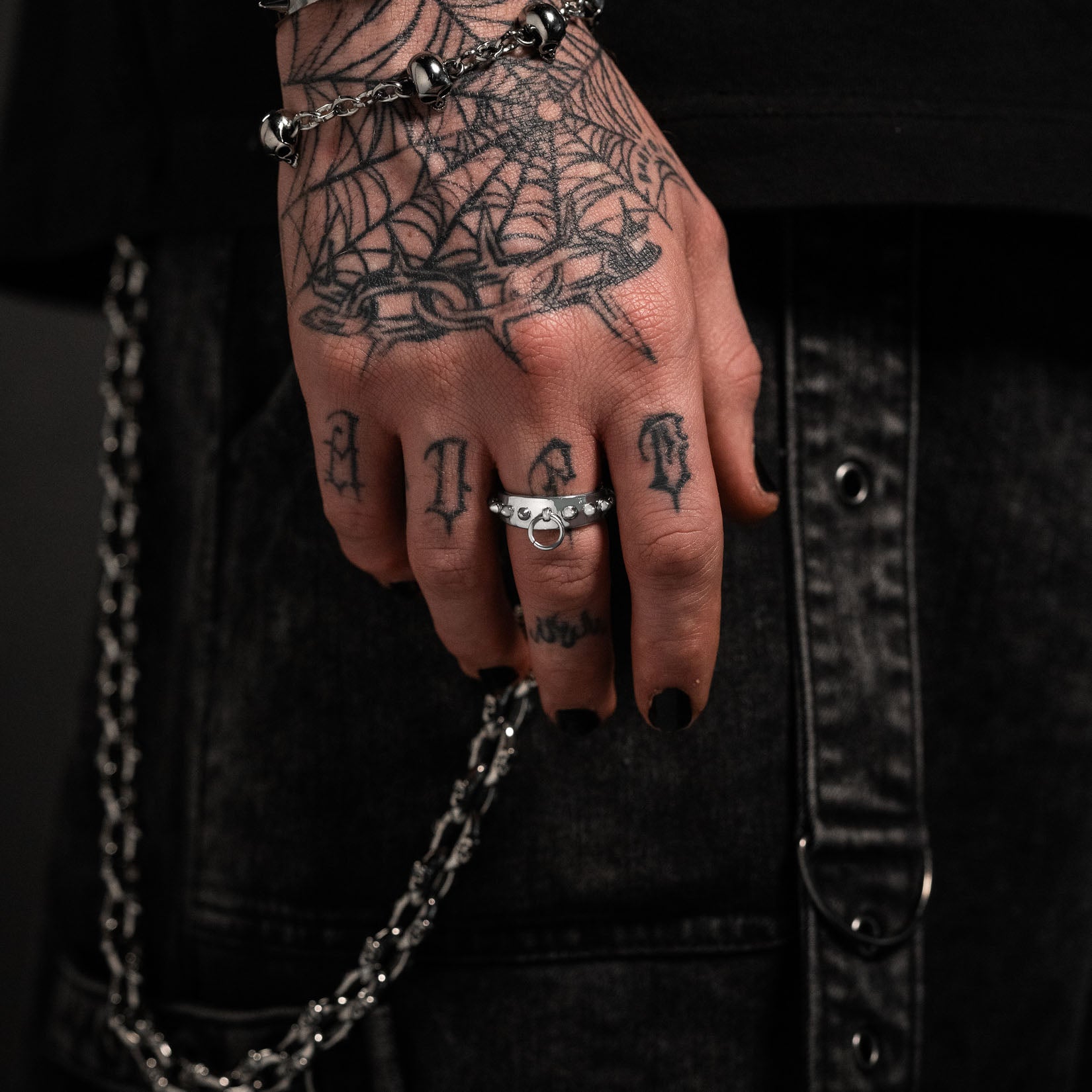 Hand with tattoos and spiked ring, wearing a chain, against a dark background by statement collective