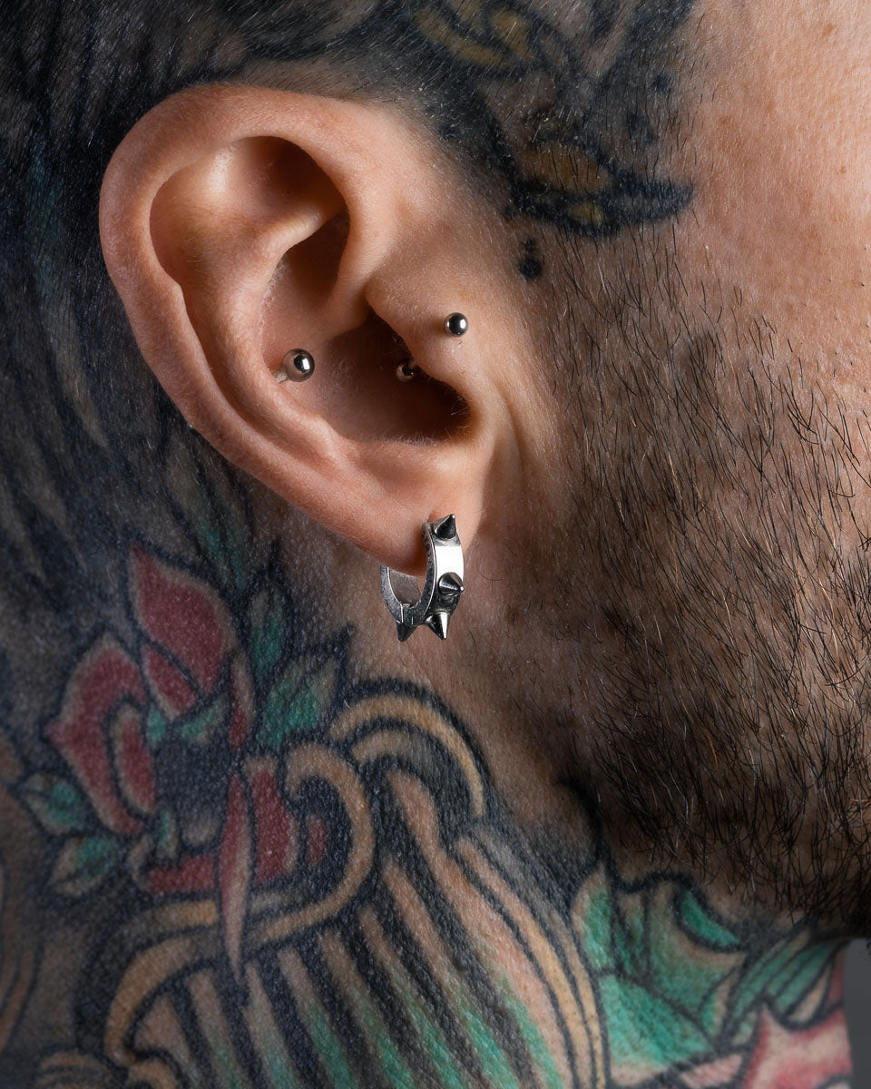 Close-up of a person’s ear with two small piercings, wearing Statement Collective 8mm Spiked Hoop Earrings. Tattoos are visible on their neck and face, with facial hair along the jawline.