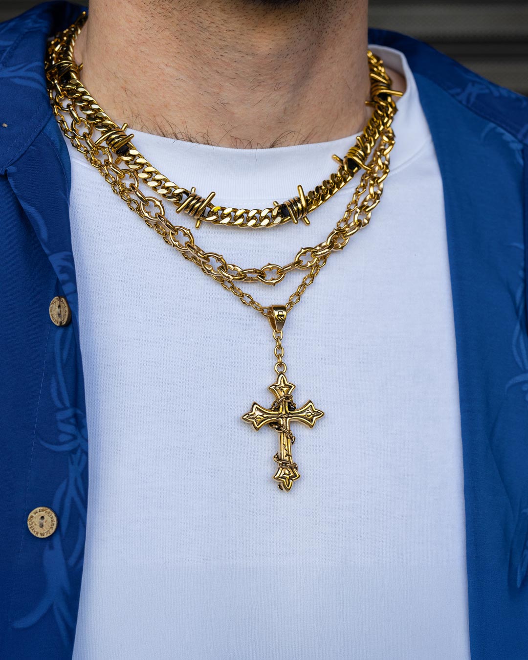 A person wears the Spite Stack Gold (Custom Pendant Choice) chain—a layered 18k gold barbed wire Cuban chain and cross pendant—over a white T-shirt and blue button-up, with only their neck and upper chest visible.